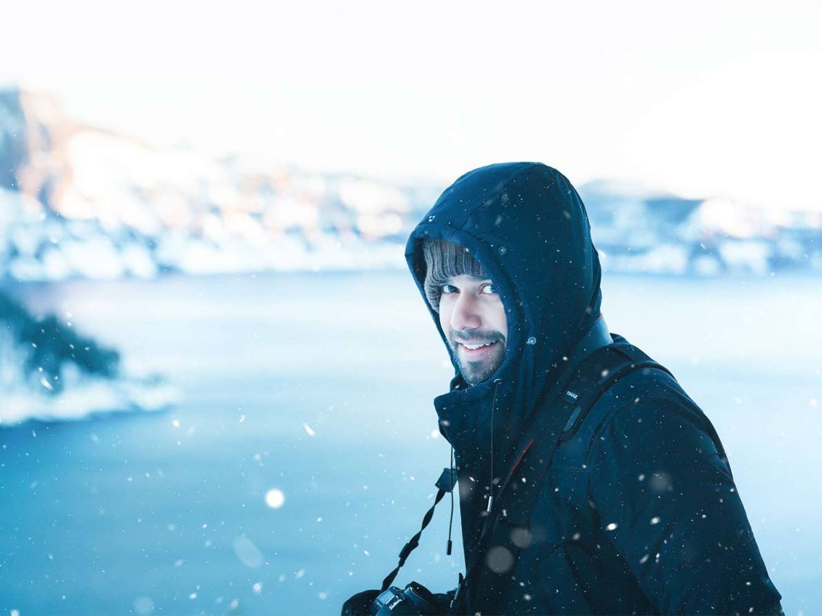 A bearded man wears a parka and stands in front of a snowy backdrop.