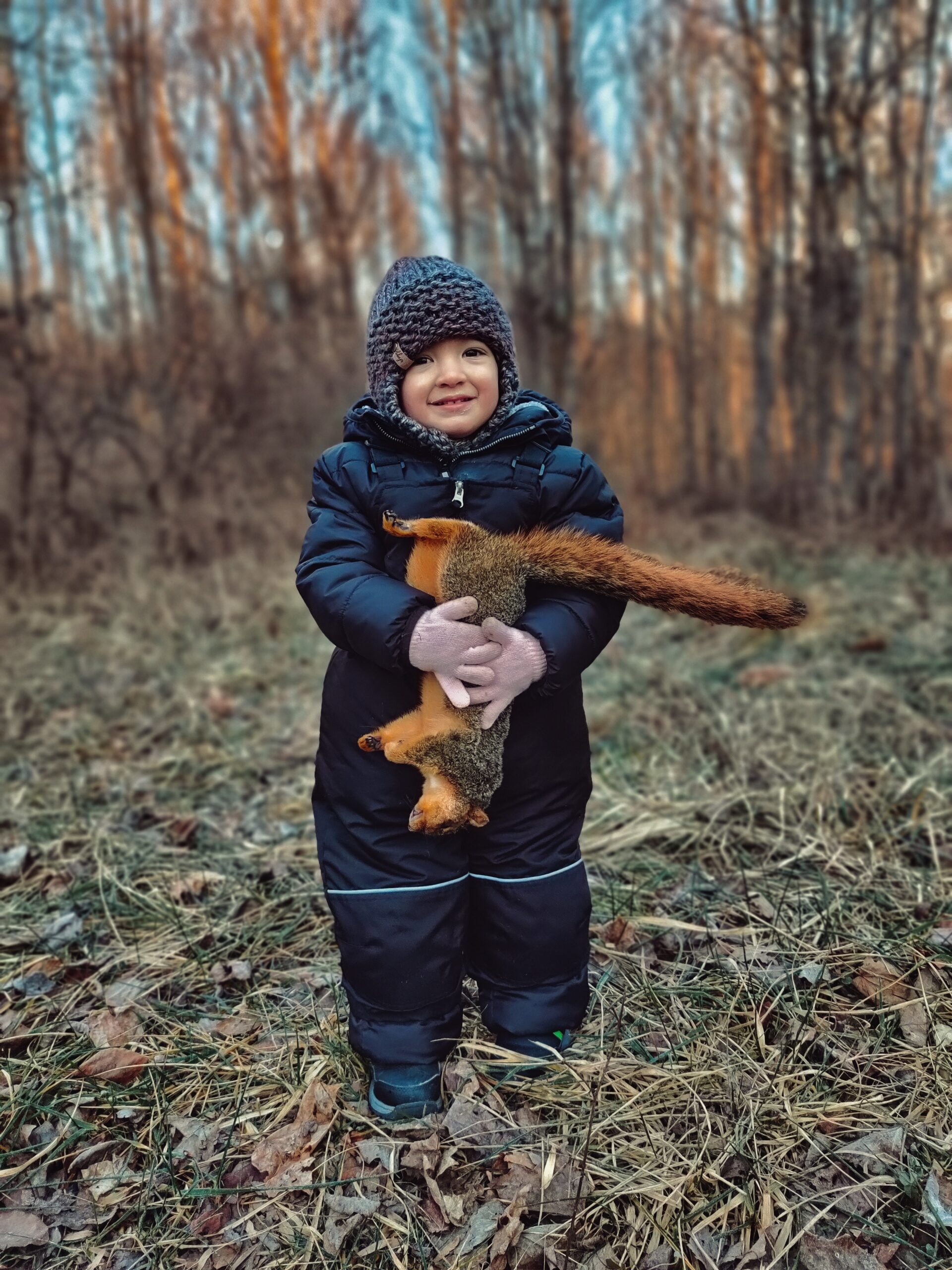 A young kid in a snow suit holds a fox squirrel and smiles.