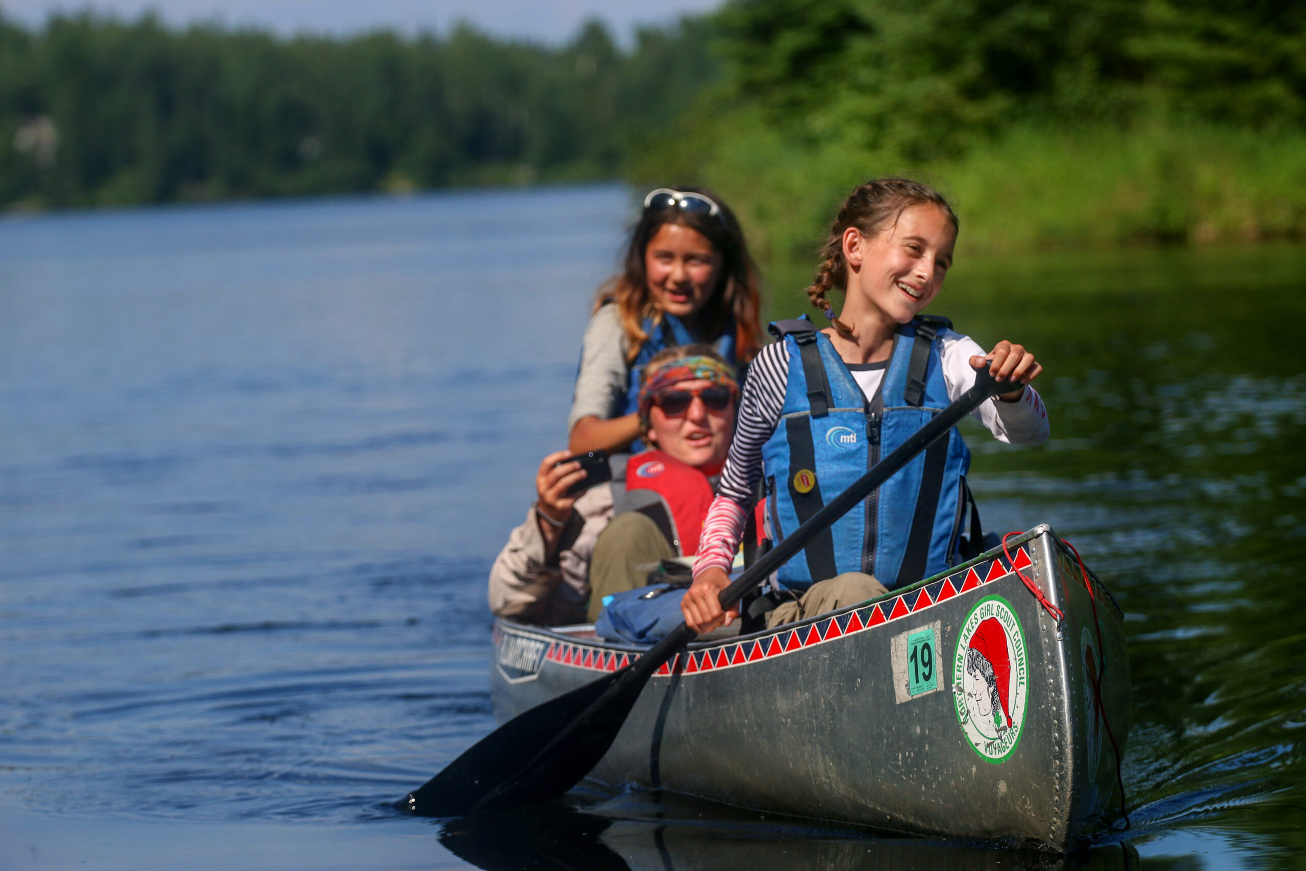 Two girls paddle a canoe on a northern like while a counselor sits in the middle of the boat.