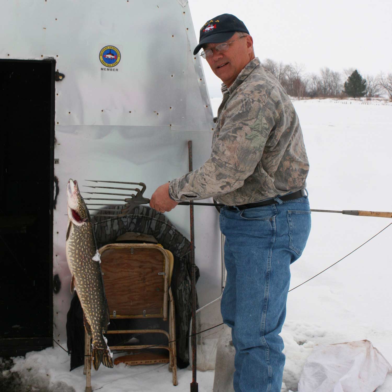 A man stands next to a large shed and an ice fishing hole.