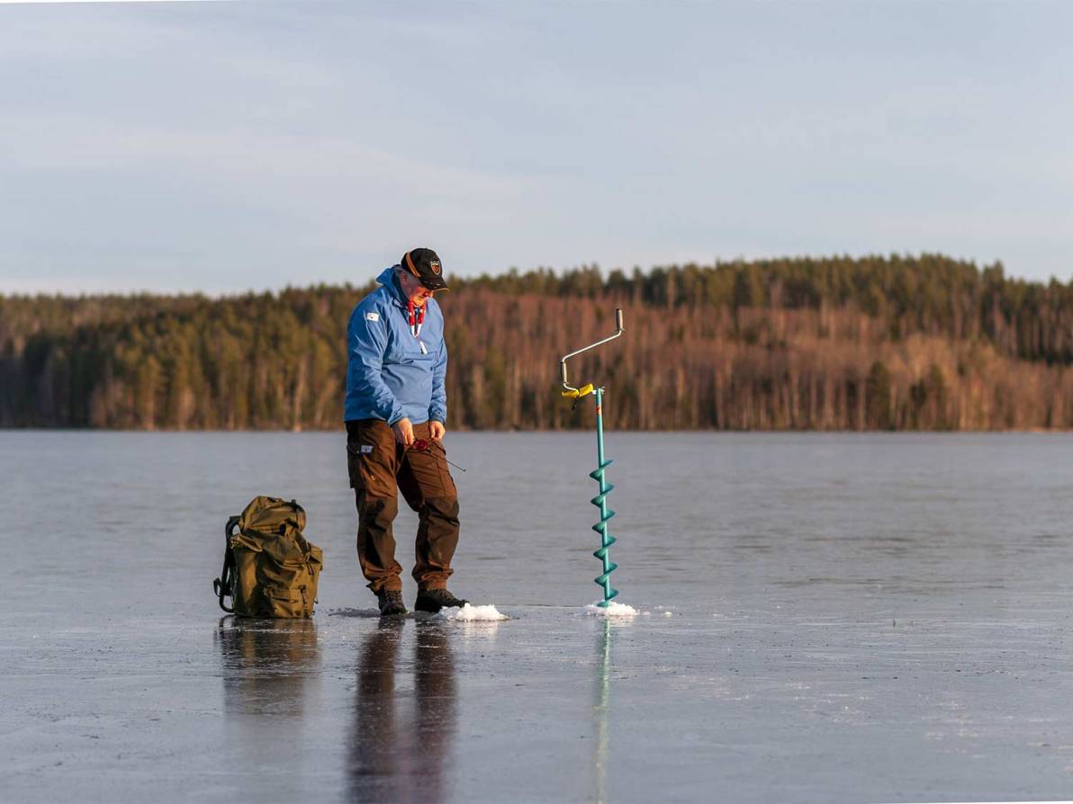 A man walks on a frozen lake, using an auger to drill a hole into the ice.