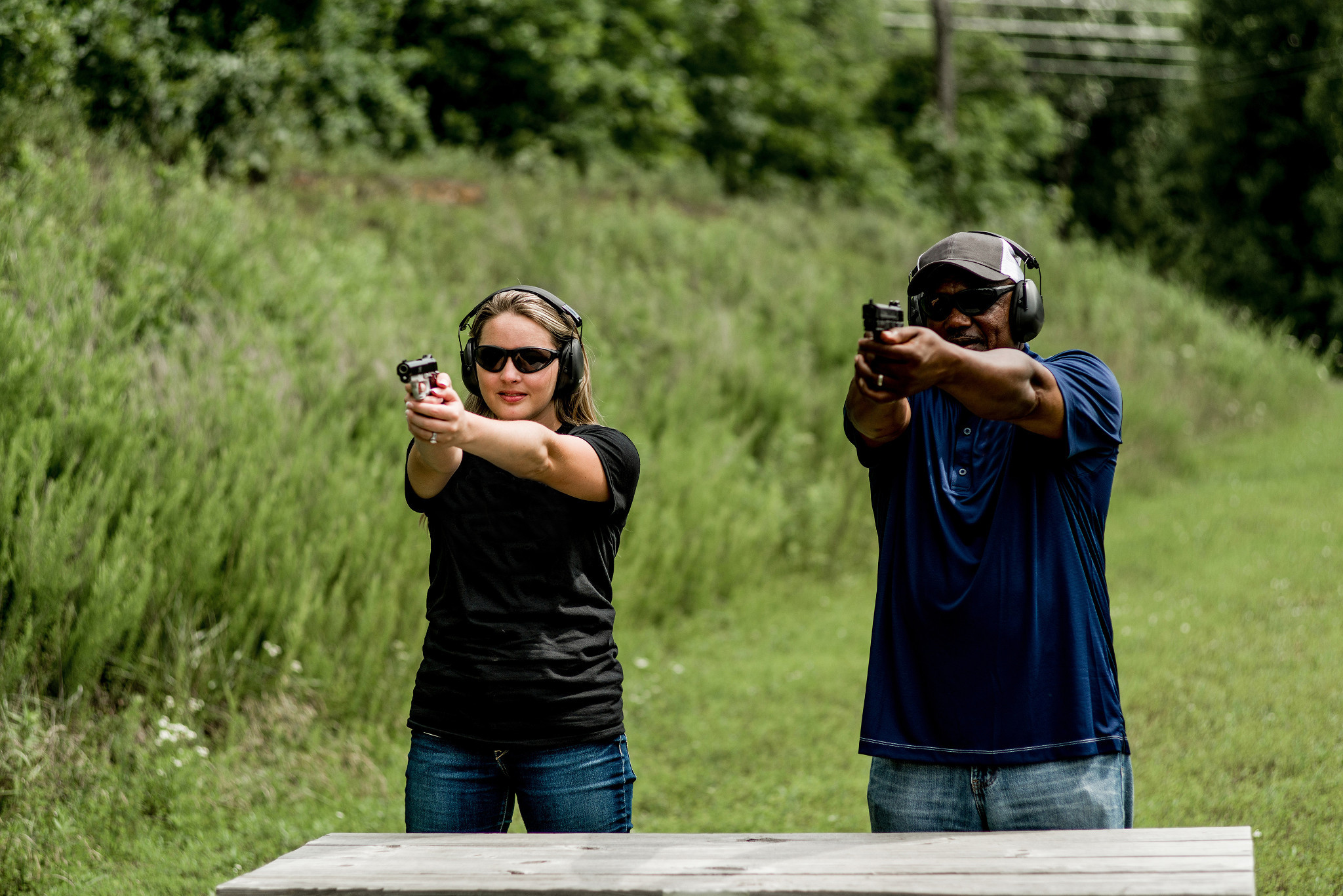 A white woman and a Black man holding handguns stand behind a table at an outdoor shooting range.