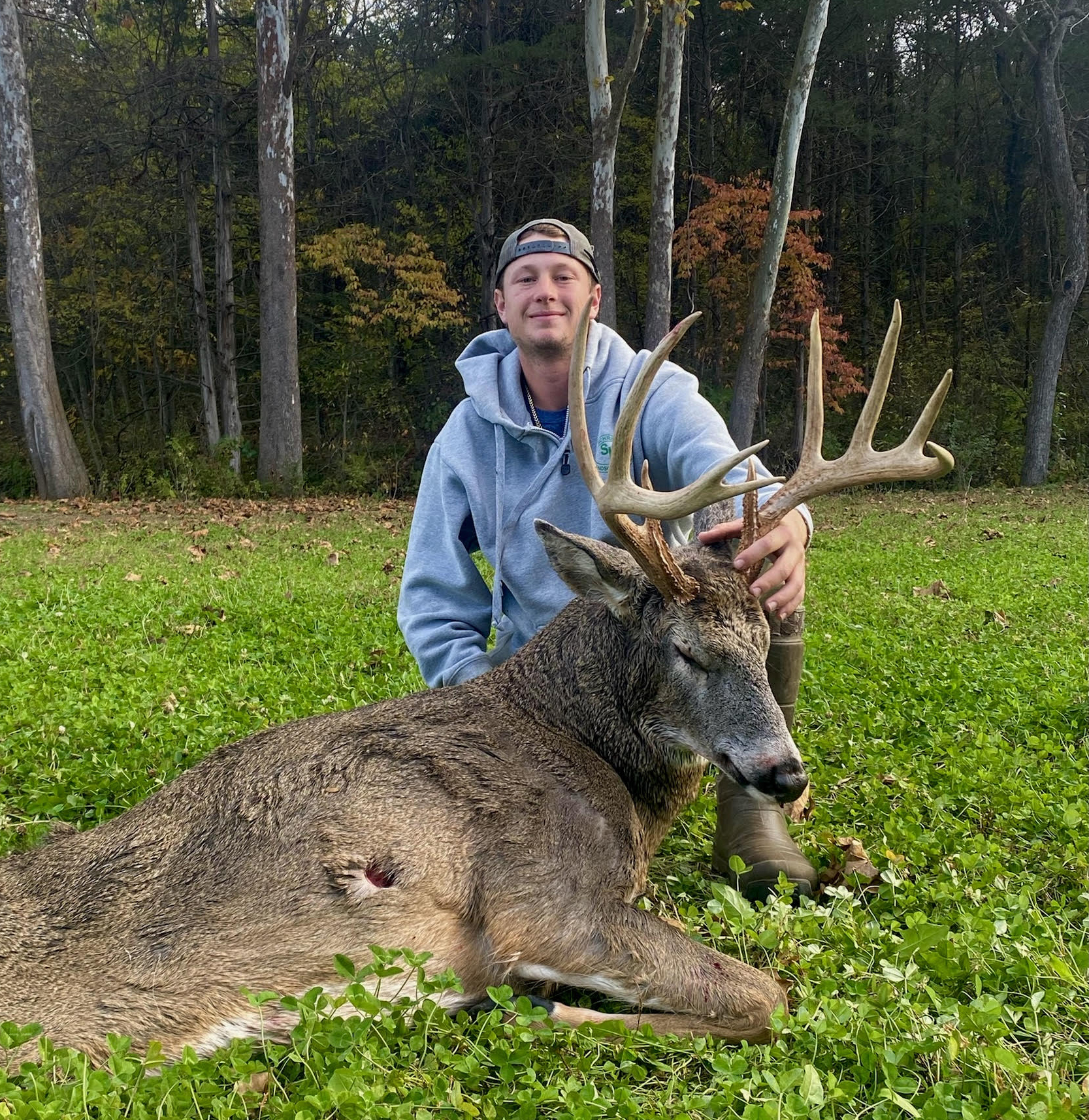 A deer hunter in a hoodie and hat smiles with his mature Missouri buck in a food plot.