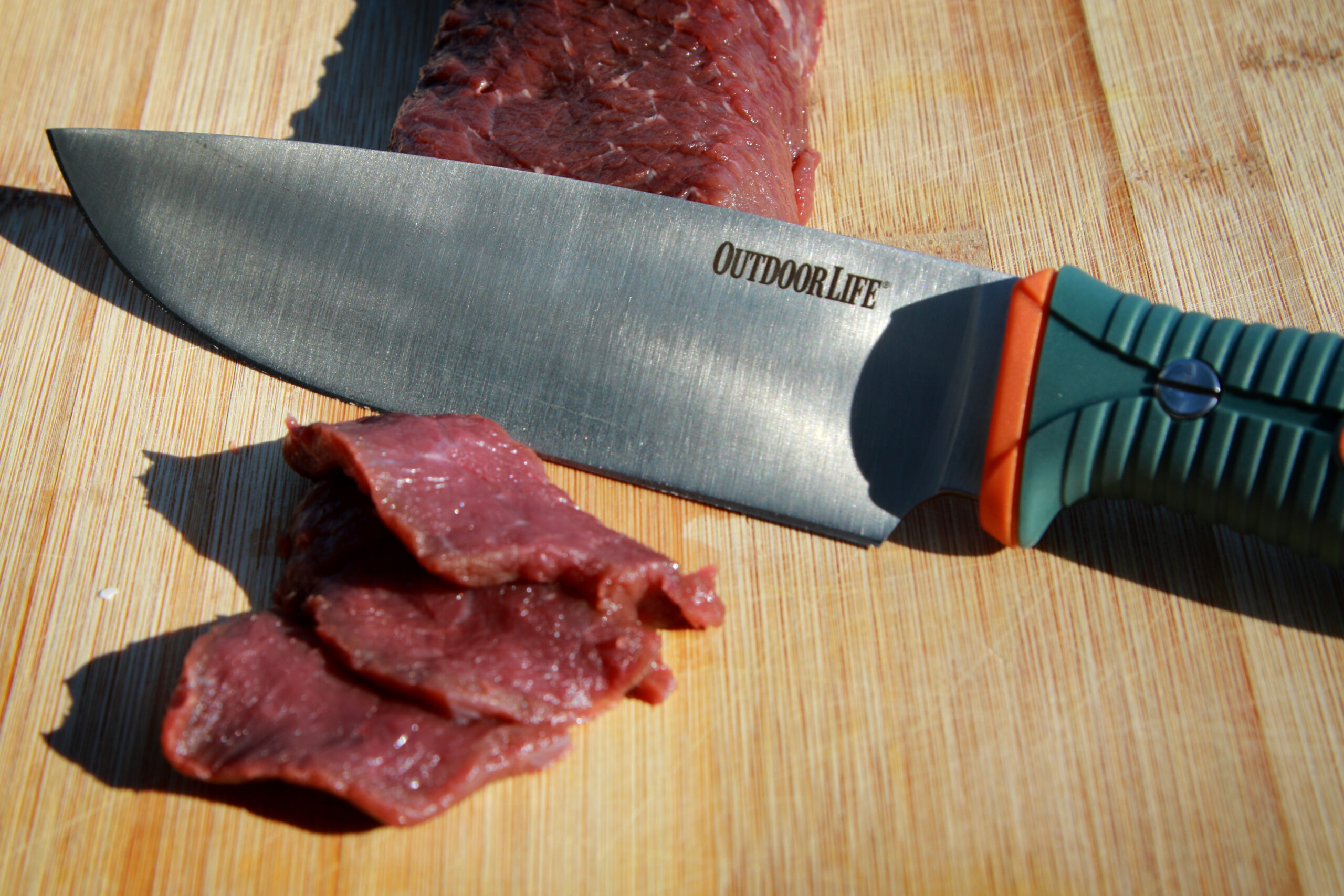 An Outdoor Life chef's knife with venison on a wooden cutting board.