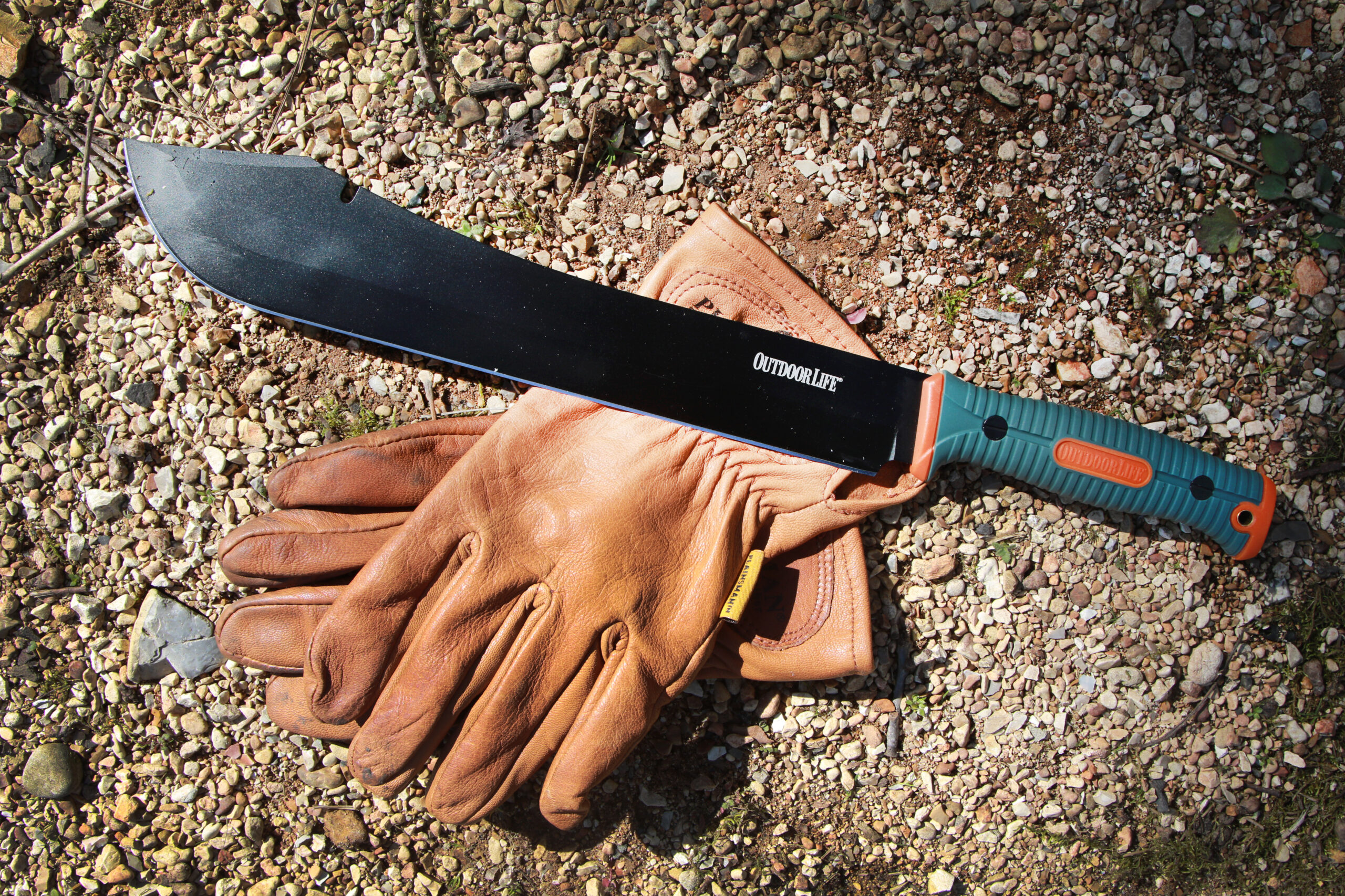 A black-blade camp machete beside a piar of gloves in the gravel.