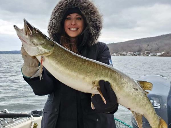 A woman smiles and poses while holding a large muskie fish.