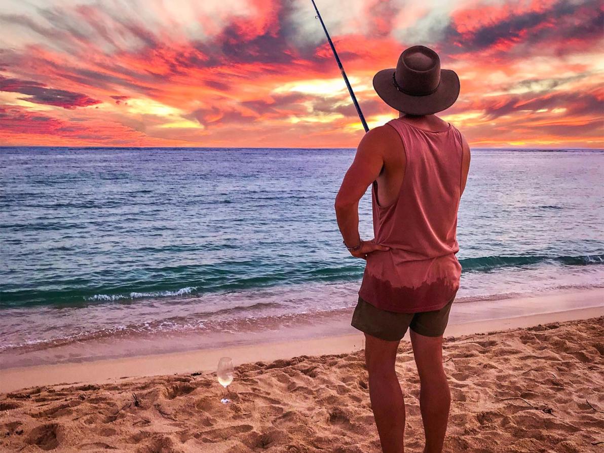 A man wearing the best fishing hat stands on the shores of the beach and fishes.