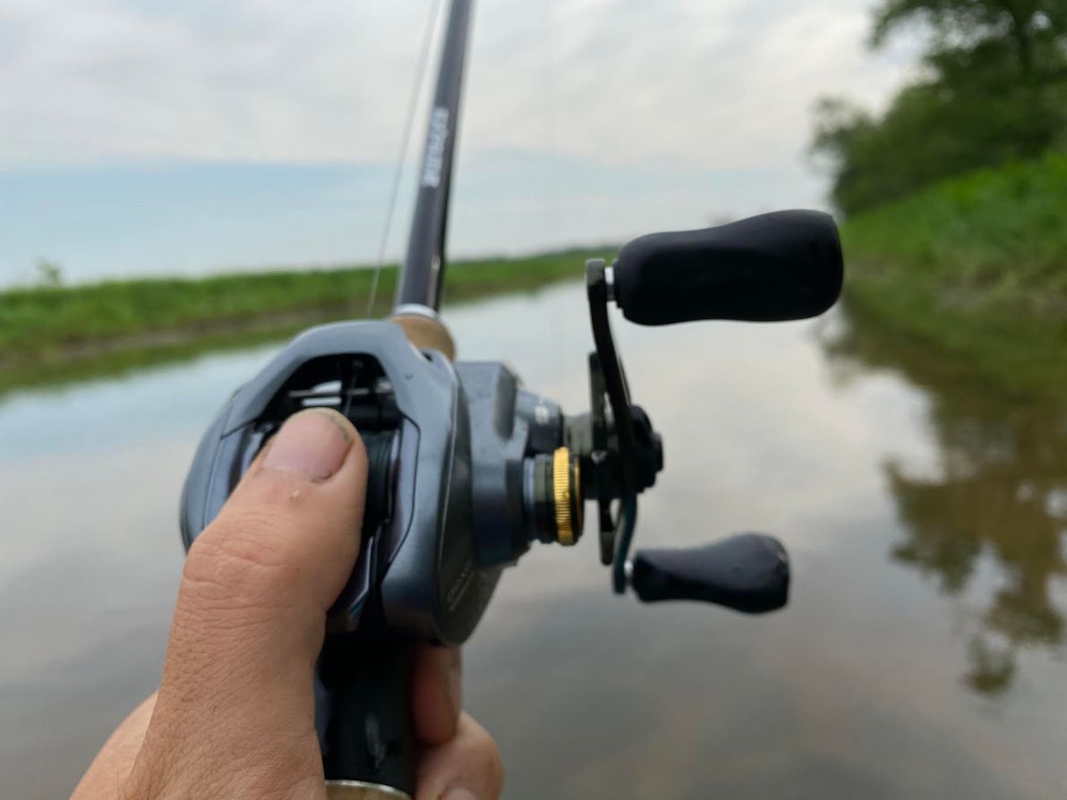 An angler's thumb on a baitcasting reel over a body of water