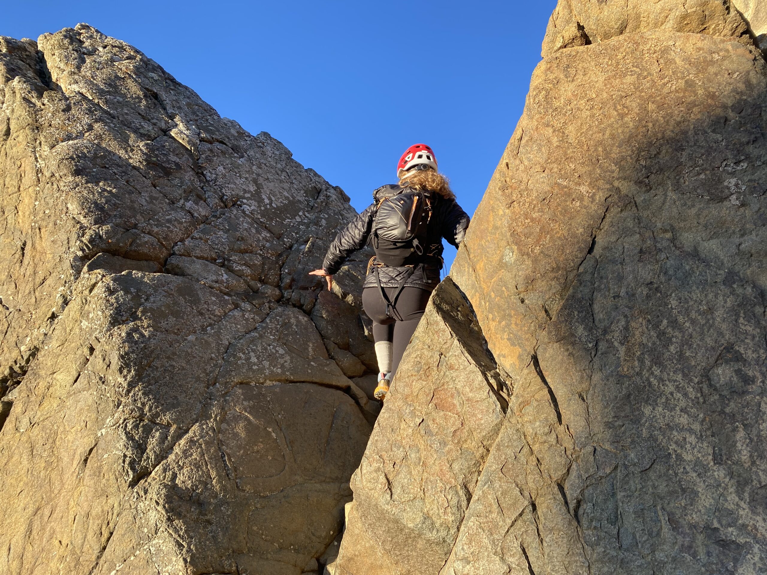 A woman with a red climbing helmet and black backpack climbing through two large rocks.