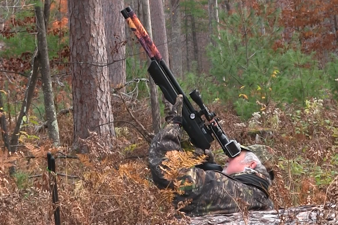 A man in camouflage in the woods aiming an air rifle up the sky