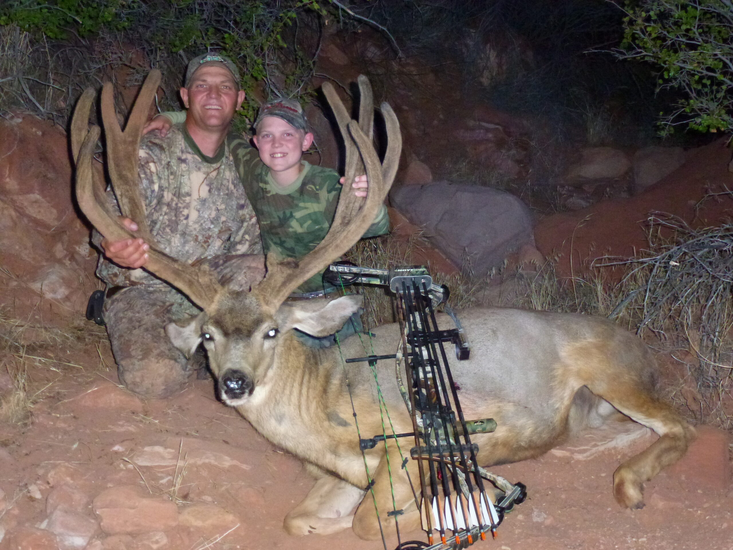 A bowhunter and a kid beside a giant velvet mule deer buck