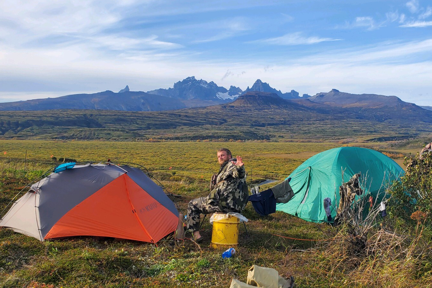 hunter sits on tundra, between two tents with mountains in distance