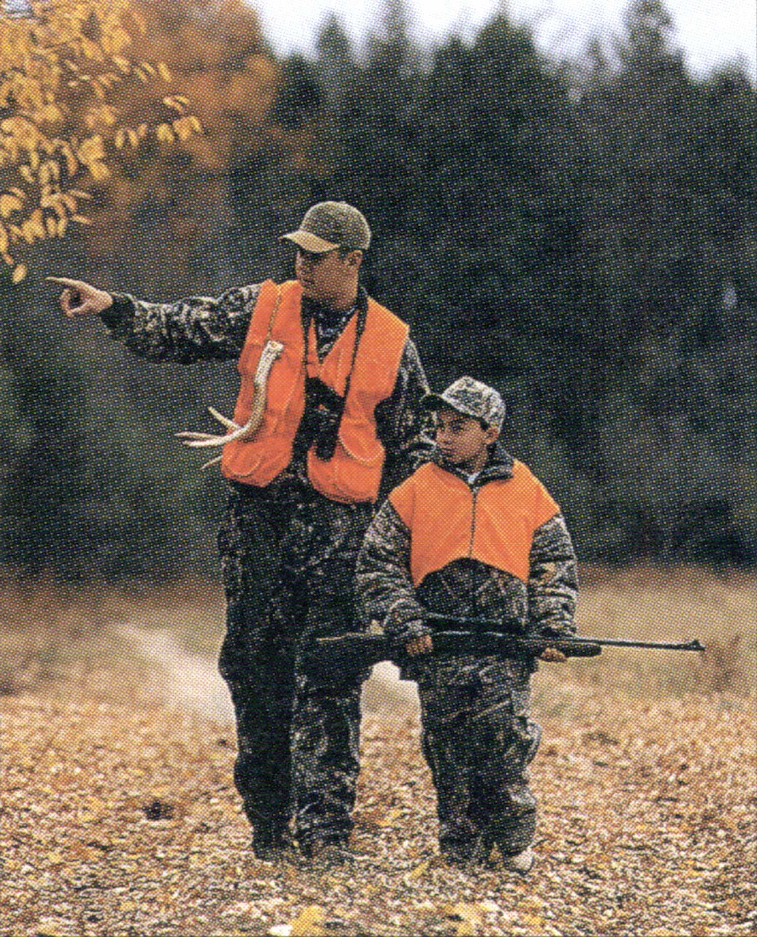 two hunters in orange vests: grown man carrying binoculars and rattling antlers walks beside young hunter with rifle