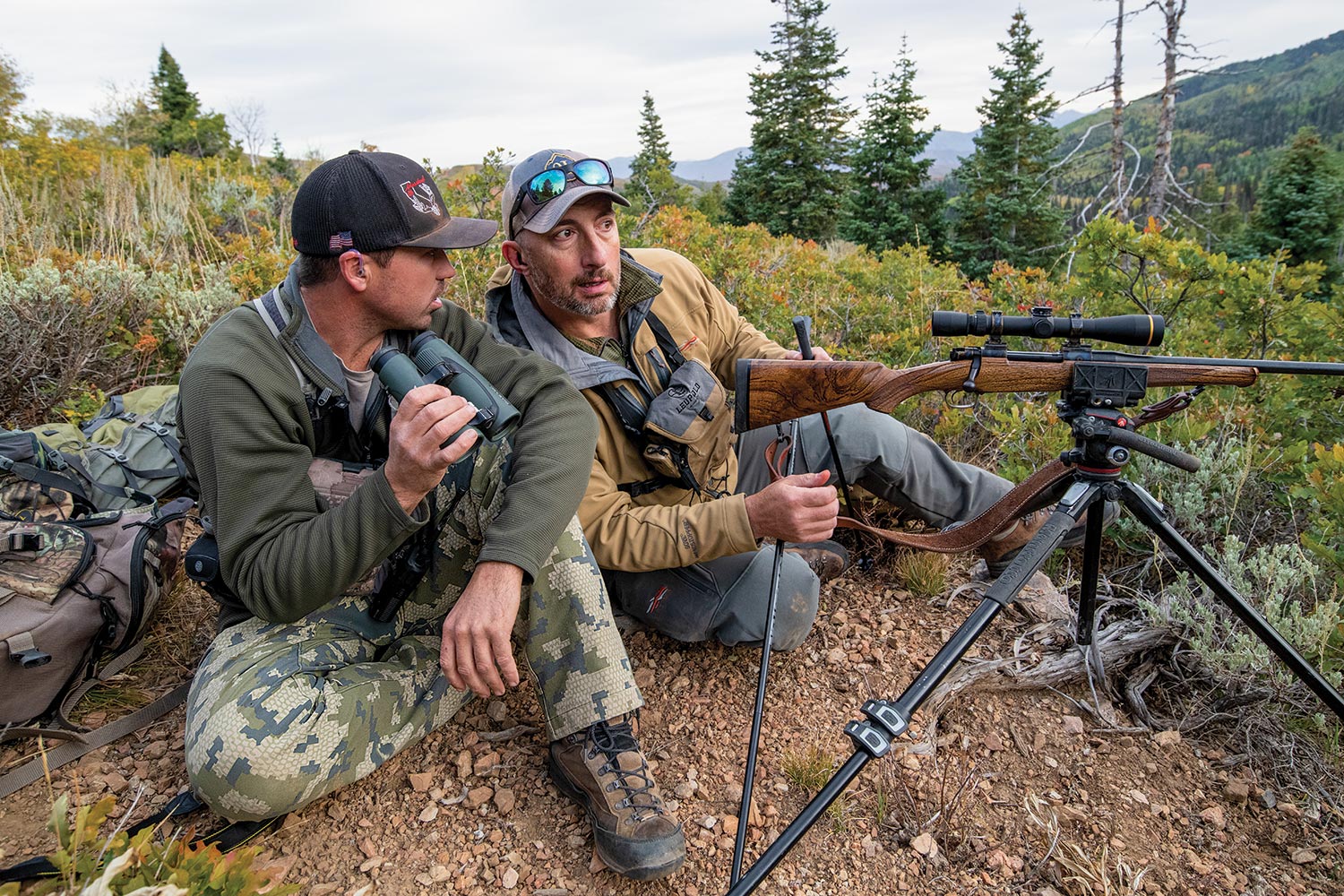 Hunter and guide, with binoculars, sit low on ground discussing plans; rifle is set up on tripod.