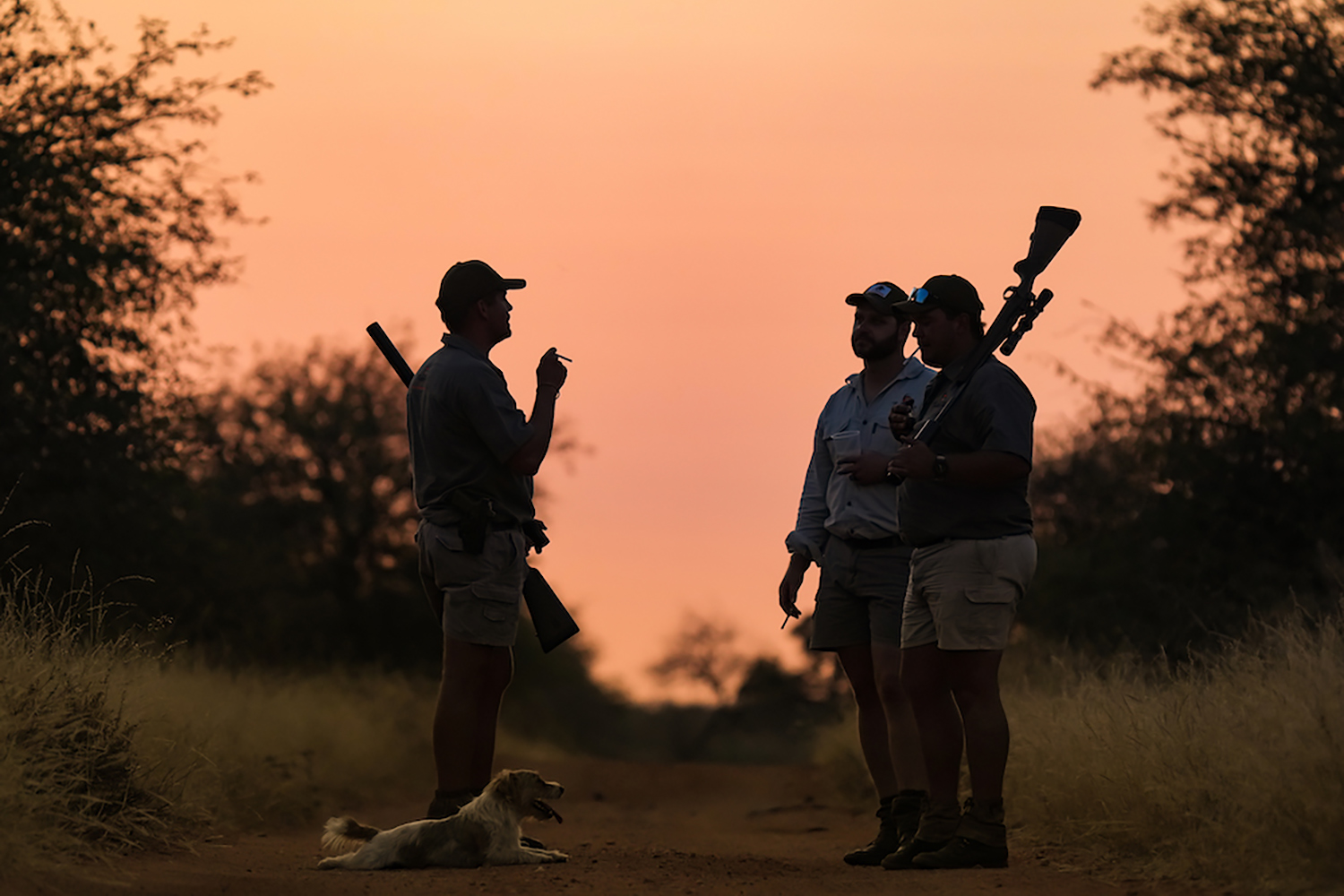 three hunters talk at sunset while dog rests at the feet of one