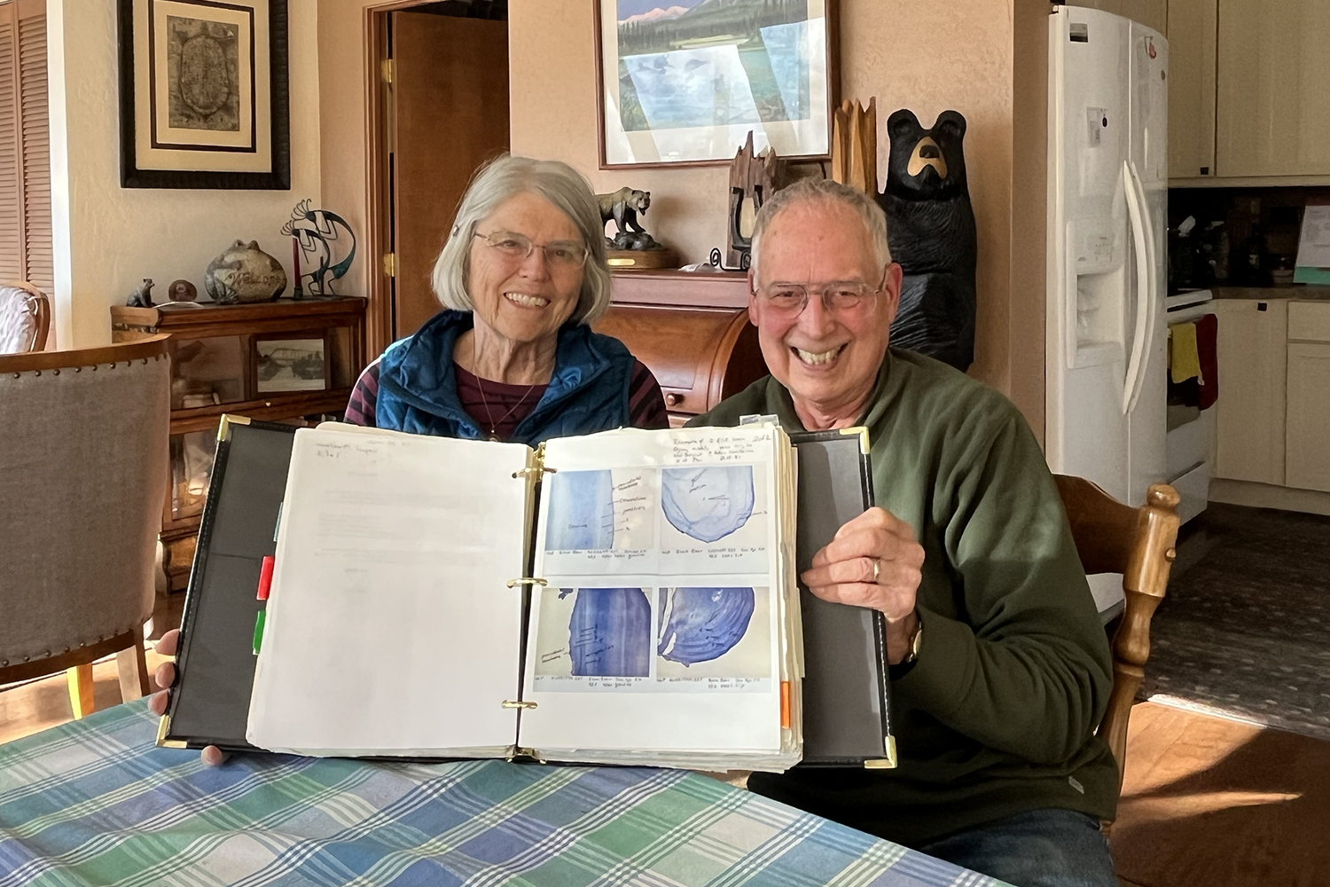 older couple sits at table and holds scrapbook open toward camera