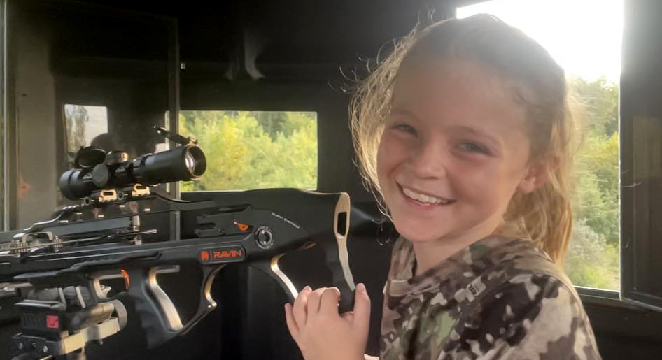A young girl sits smiling behind a Ravin crossbow in a deer blind.
