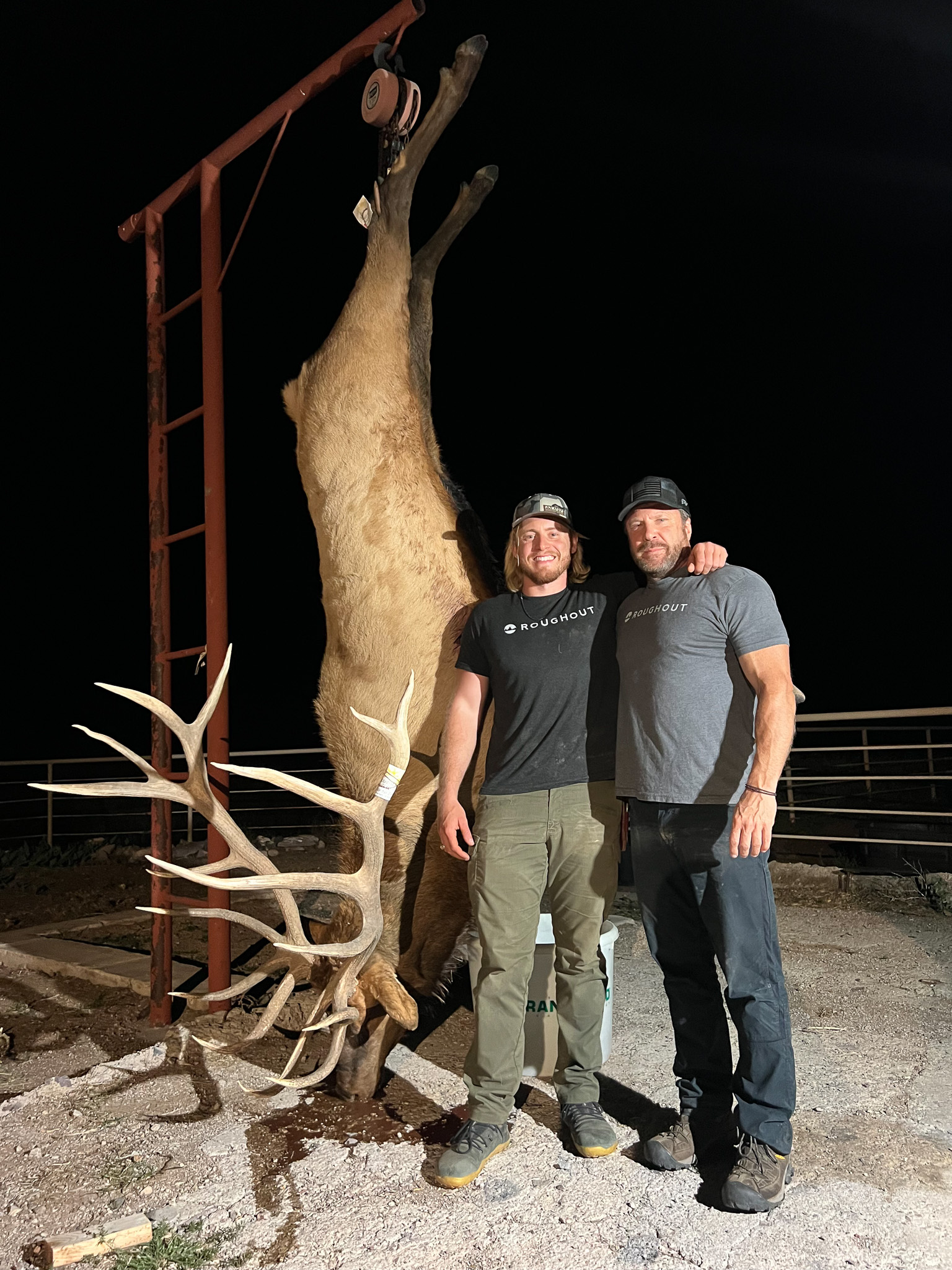 A big elk hanging from a meat pole with two hunters beside it.