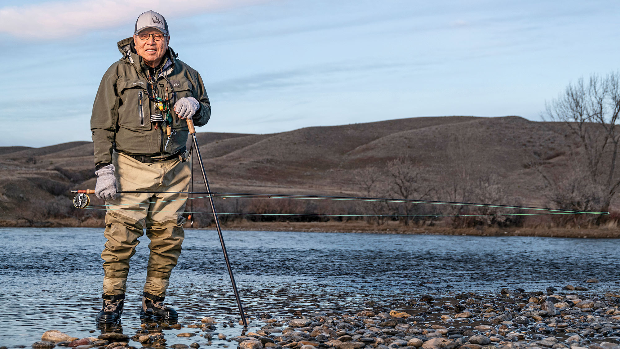 fly fisherman wearing waders and holding rod and walking stick poses at shallow, rocky edge of river