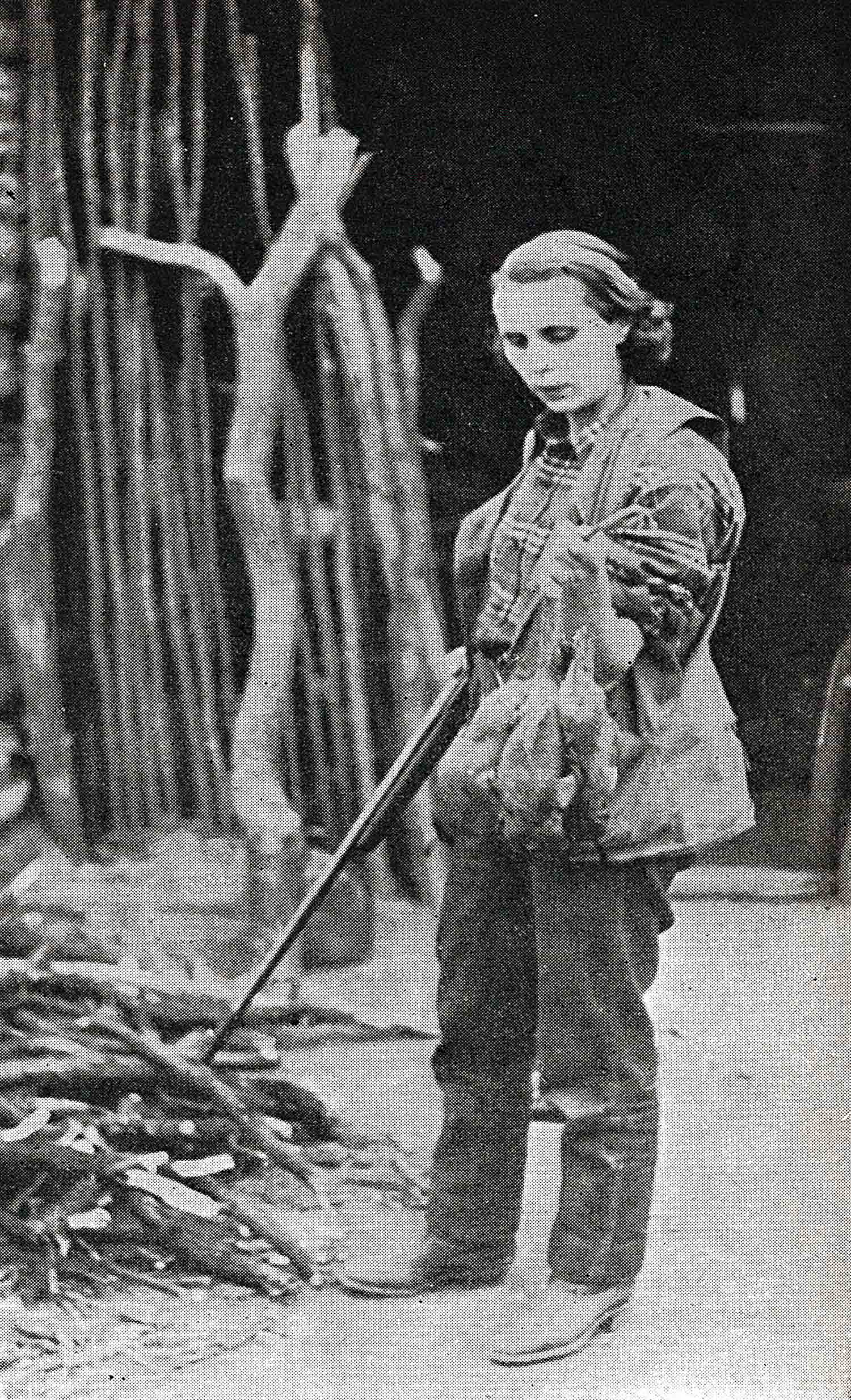 Hunter standing in front of wood pile holds long gun looks down at the quail she has shot. Archival photo.