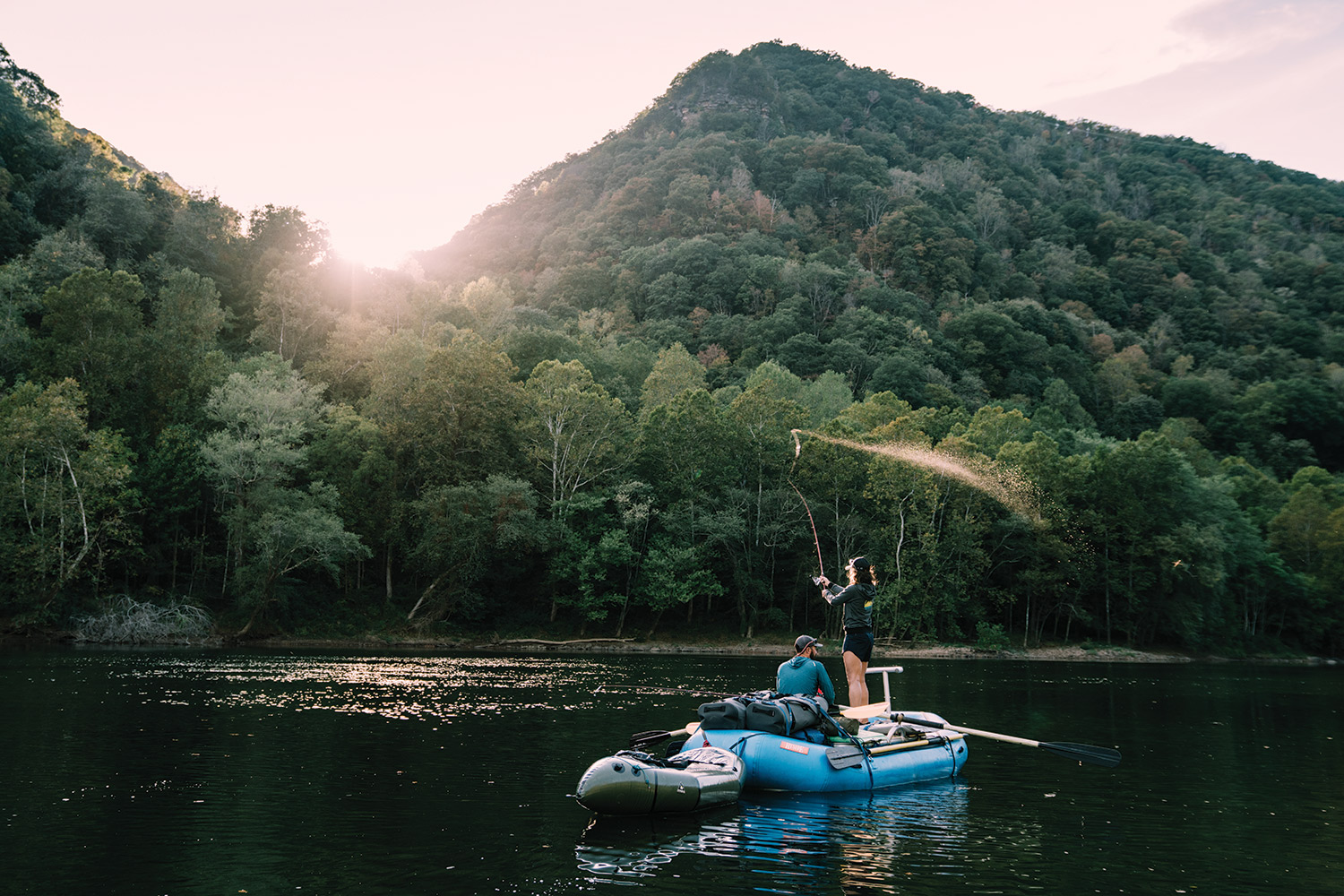 Angler stands on large raft to cast as sun sets behind tree-covered hills.