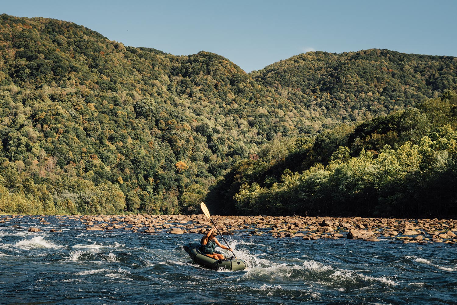 paddler in small packraft on rocky, swiftly moving river water with shrub-covered hills behind