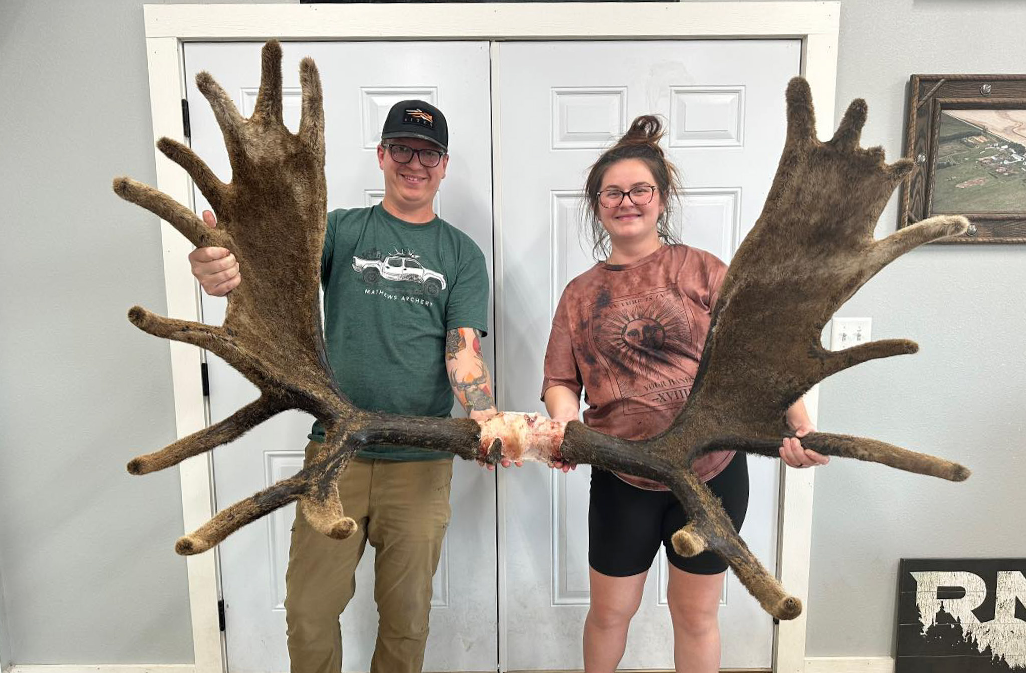 A couple holds up velvet moose antlers in front of a white closet.