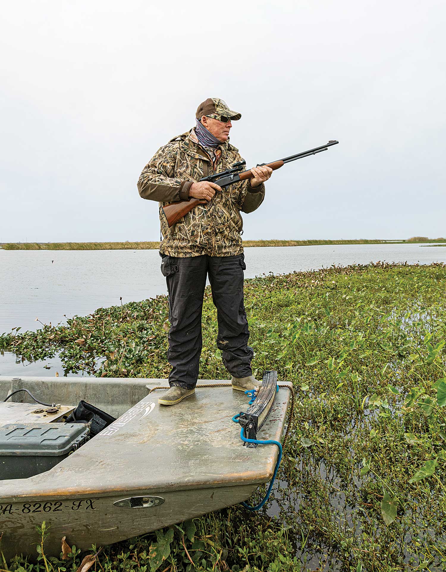 hunter holding rifle stands on boat stern in marshy area