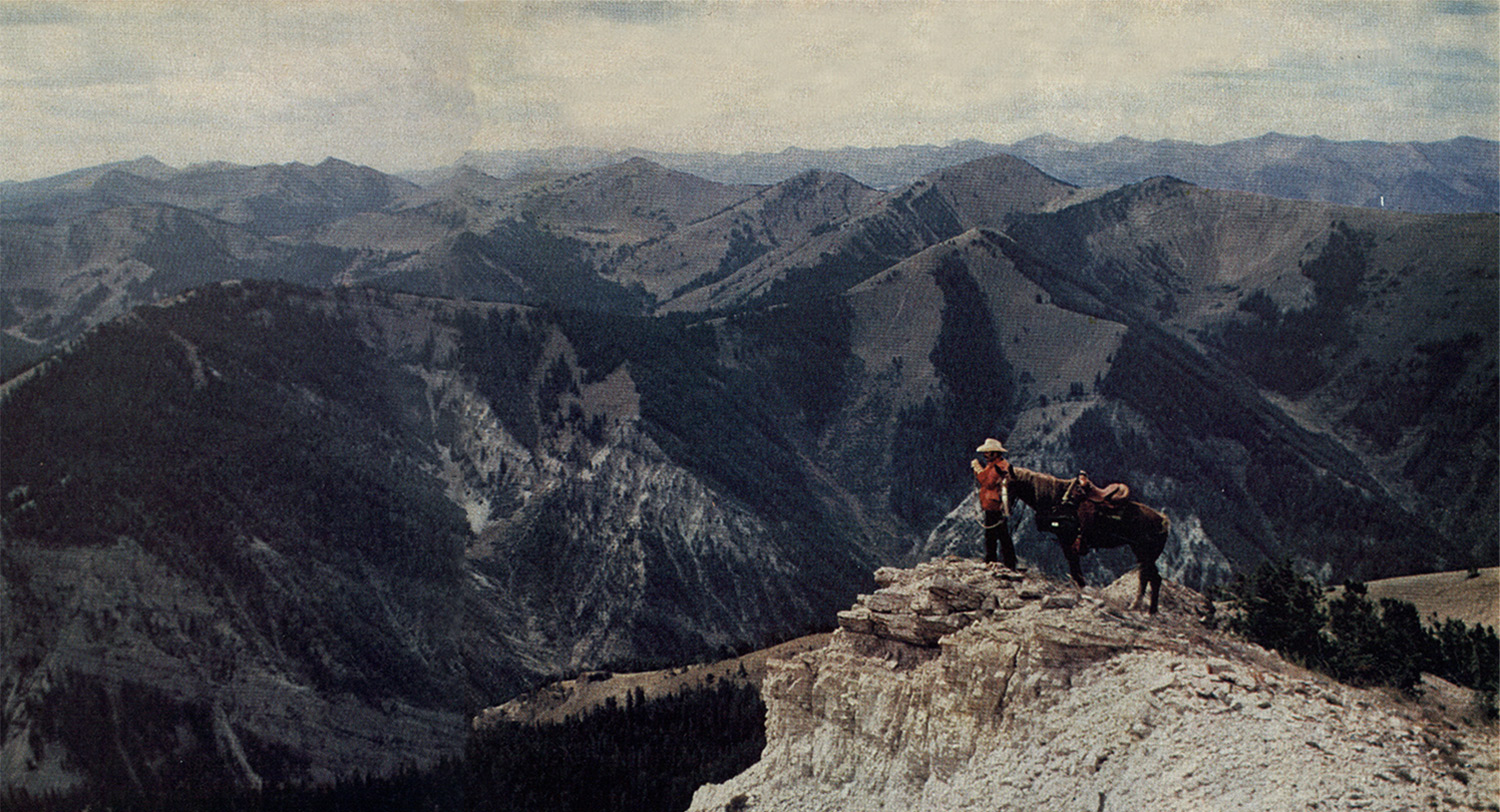 hunter looks for game as he stands on the edge of a high-elevation cliff with horse behind him; mountains in the distance
