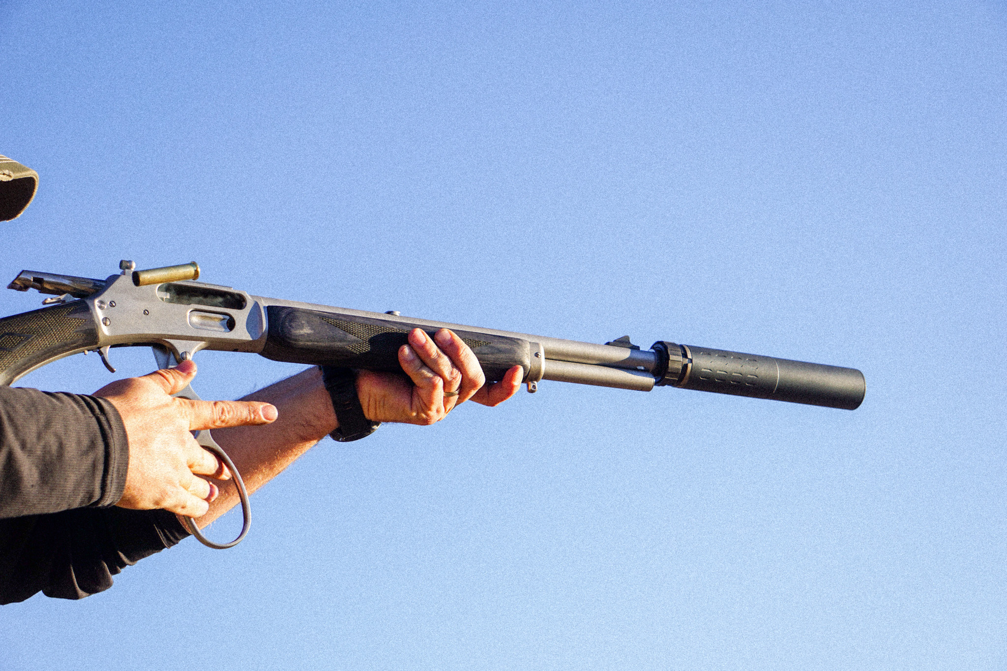 A shooter works the action on a suppressed Marlin lever-action rifle against a blue sky.