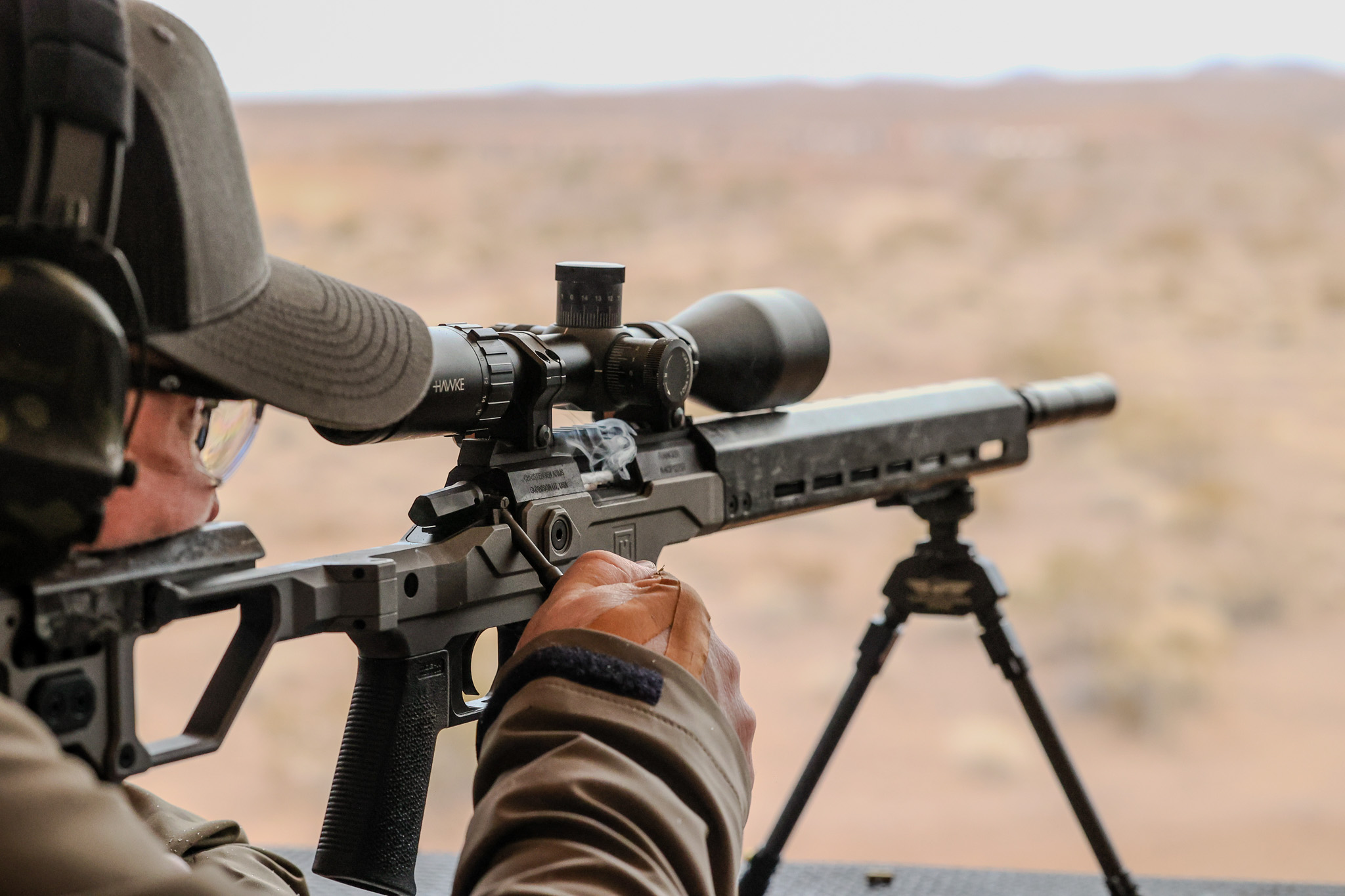 A shooter works the bolt on the action of a chassis-style bolt-action rifle on a bipod at a desert range.