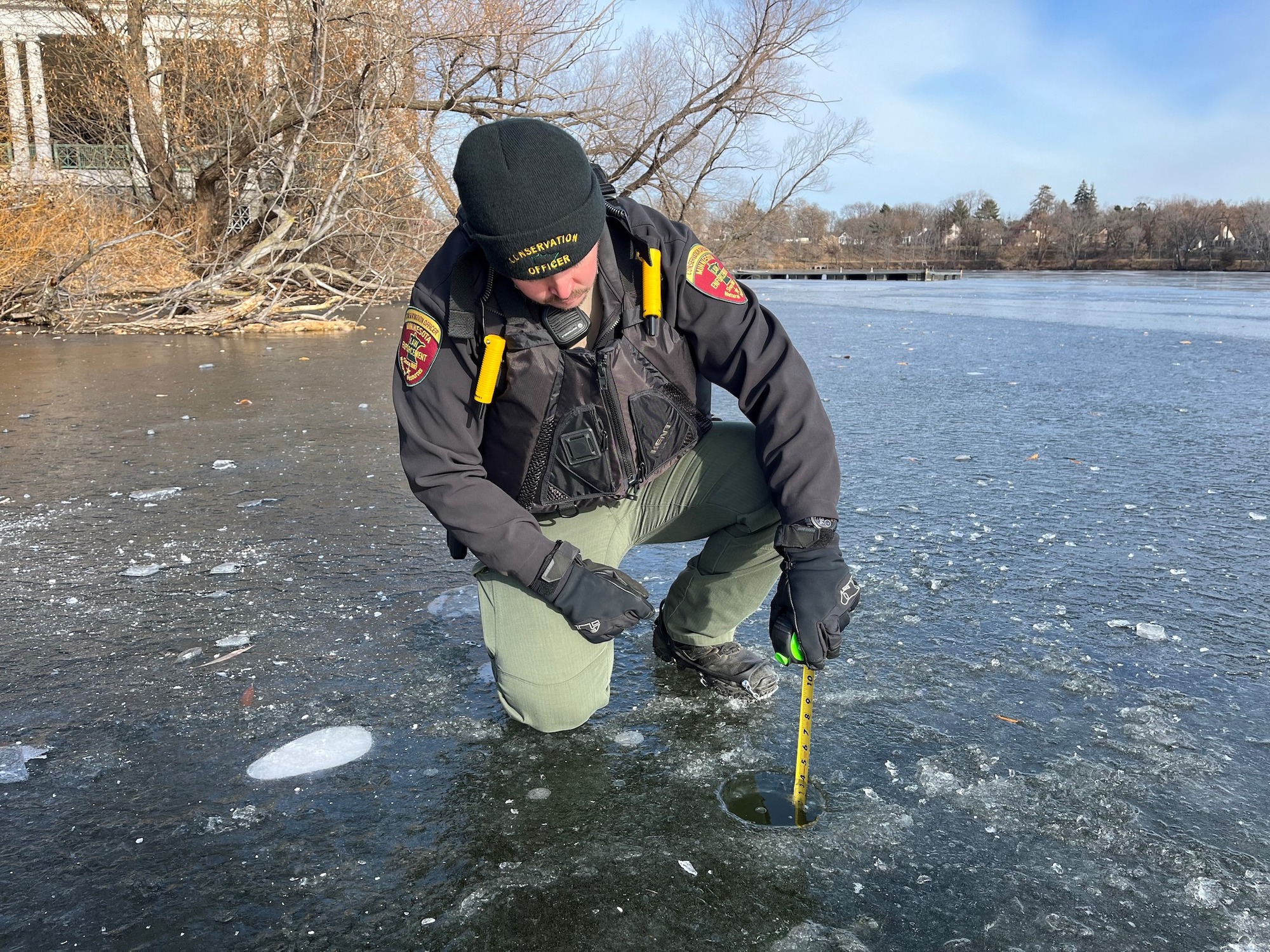 A conservation officer kneels on a frozen lake to measure the thickness of the ice.