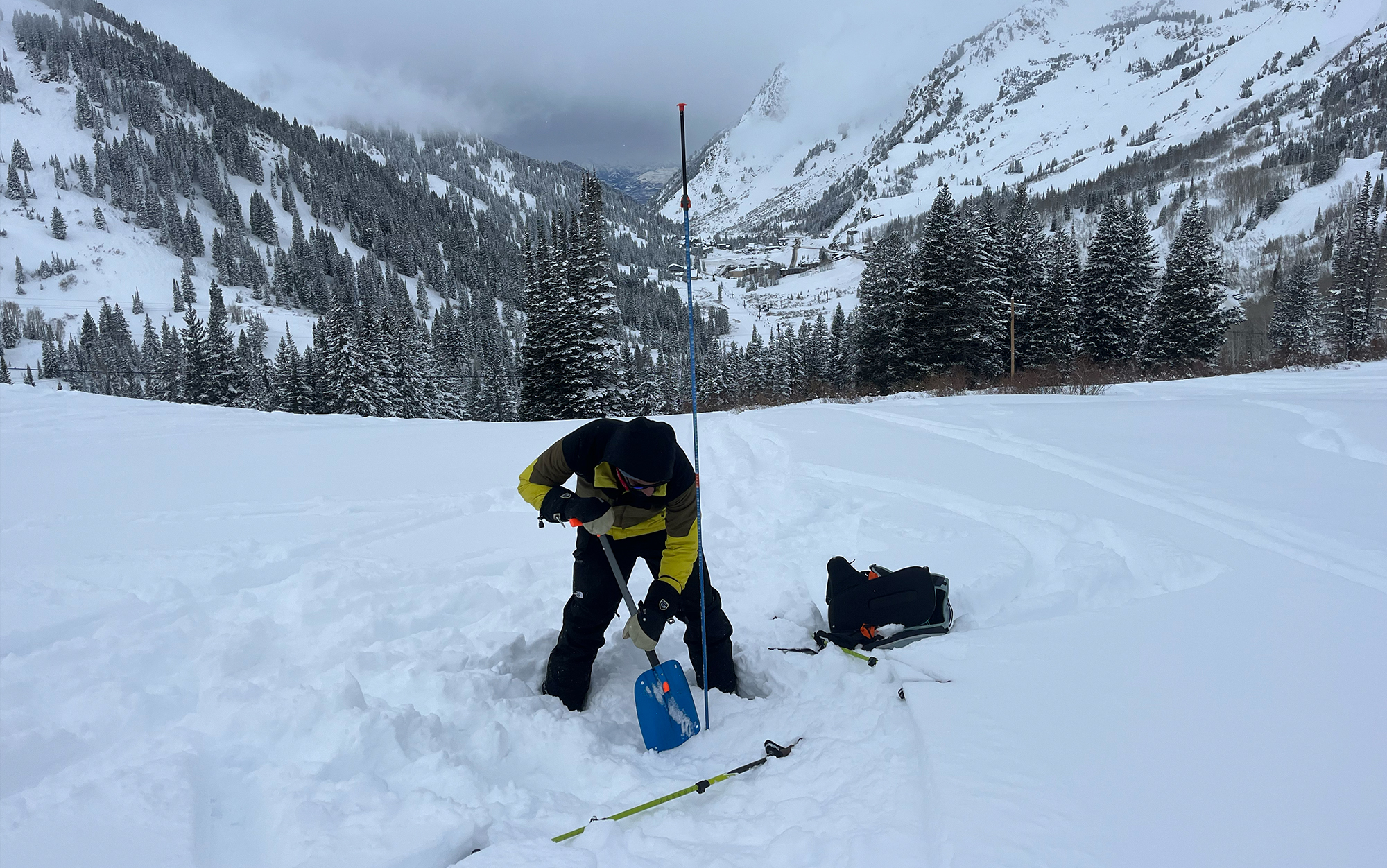 A classmate demonstrates a mock rescue scenario where he has already pinpointed the victim (an extra beacon buried in the snow) with a probe and begins digging.