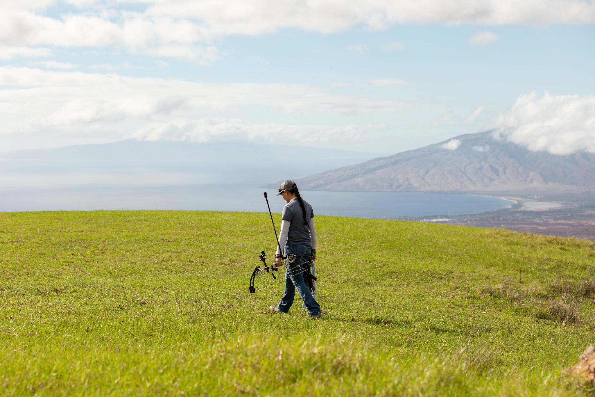 Liko Arreola walking with her bow in a grassy field overlooking the ocean on Maui.