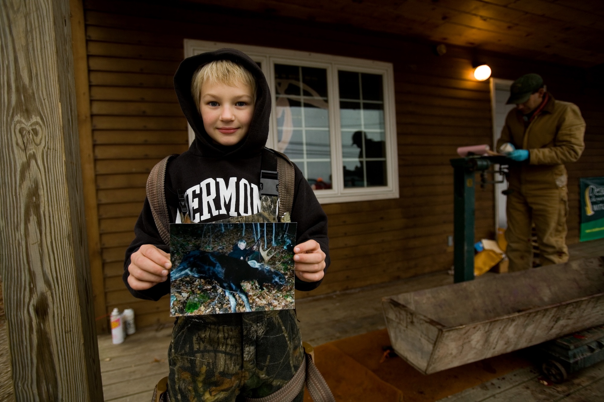 A young Vermont hunter holds up a photo of a moose he killed while a wildlife biologist examines his dad's deer.