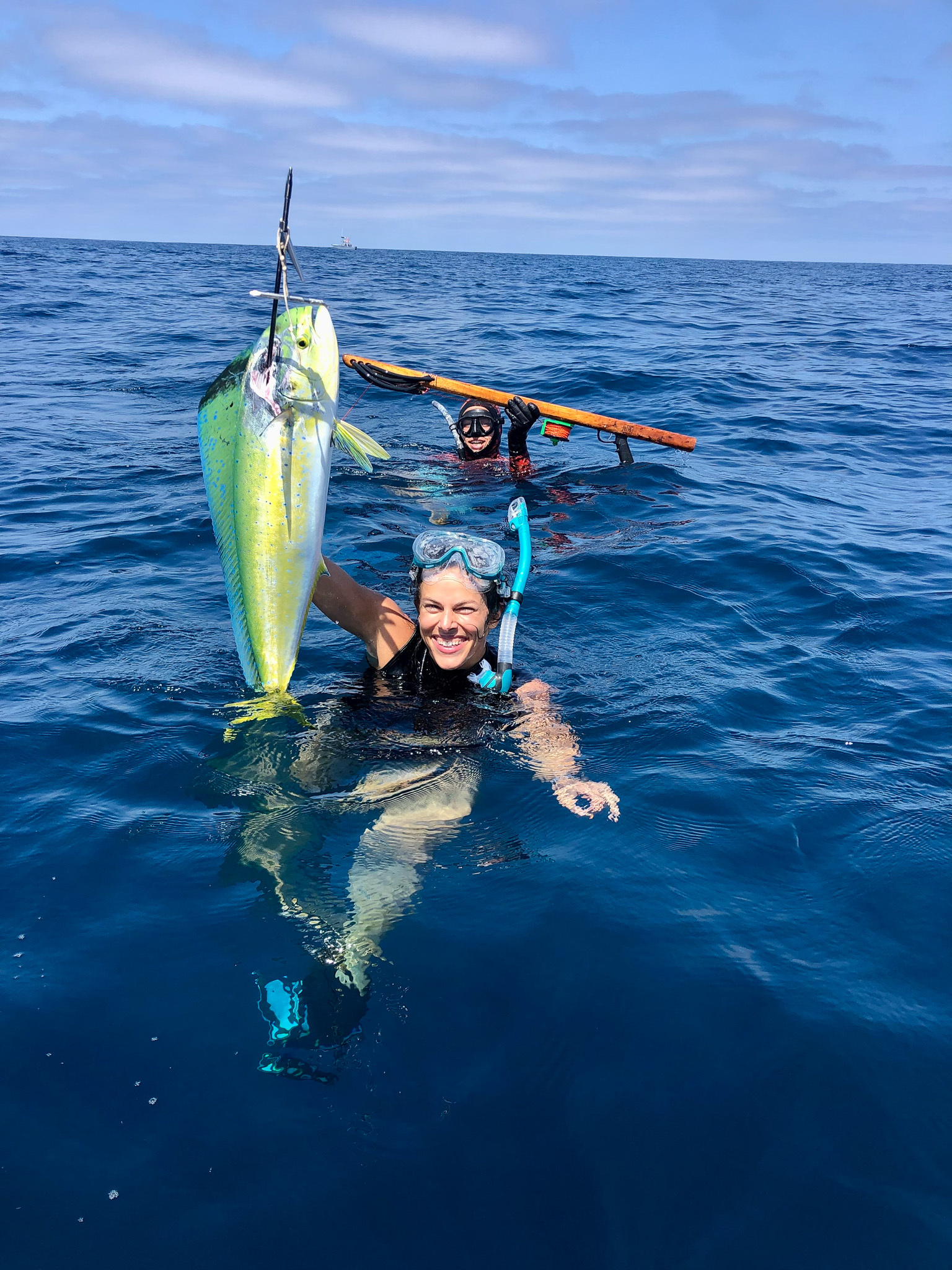 A spearfisherman holds up a dorado while her buddy holds up a speargun.