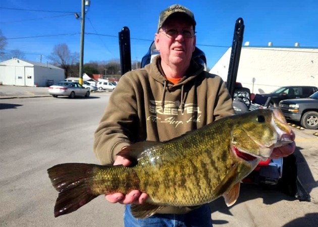 An angler holds up a smallmouth bass in a parking lot.