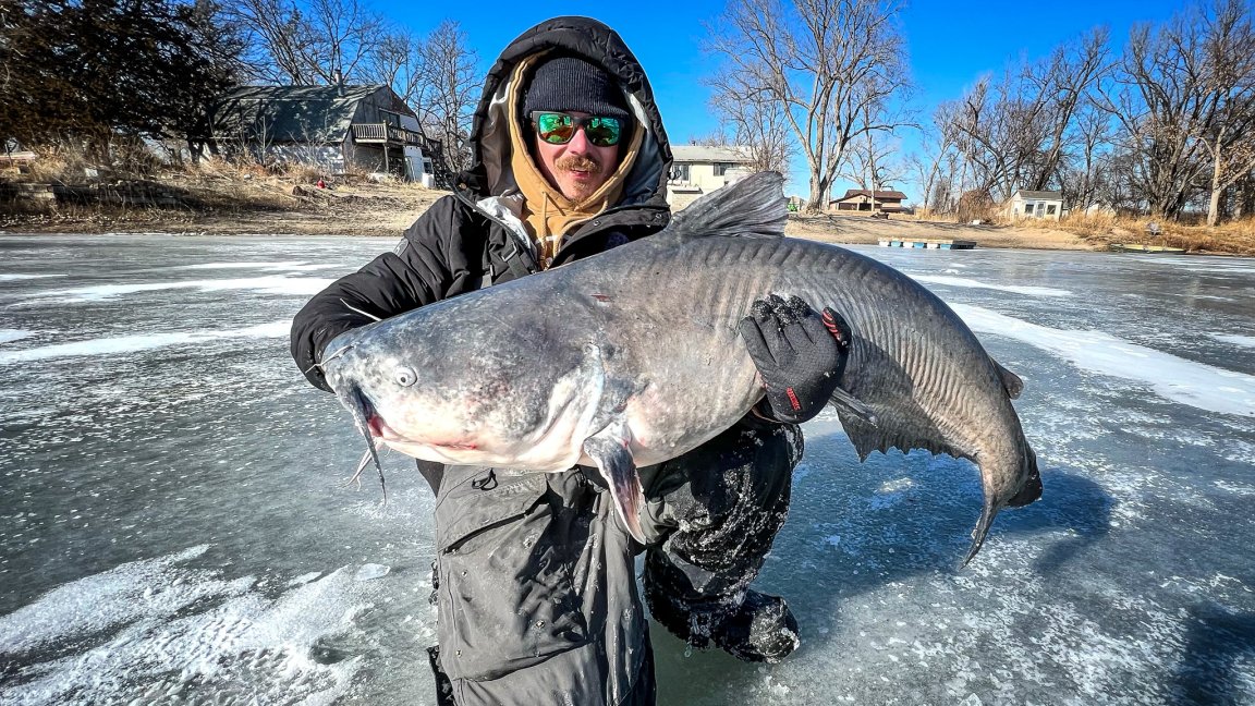 An ice fisherman kneels with a fat catfish on the ice.