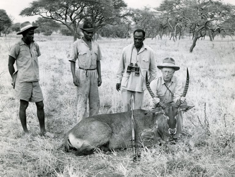 Jack O'Connor on safari, kneeling behind a waterbuck with three guides/trackers beside him.