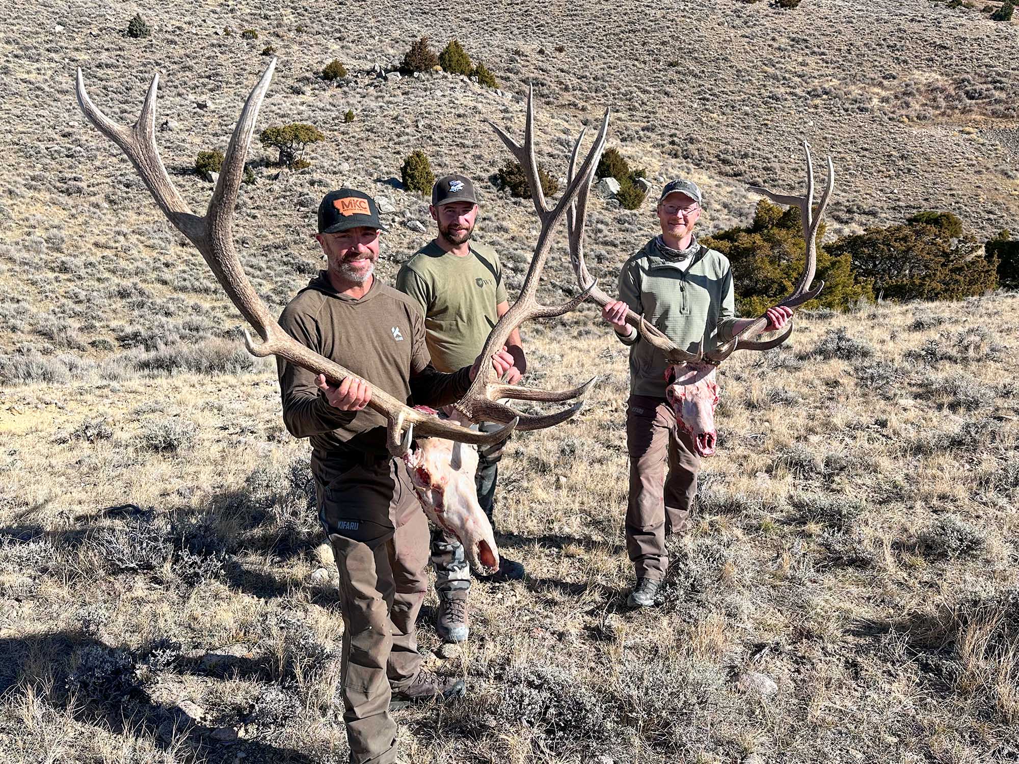 Three men stand on a Western landscape, two of them holding bull elk skulls.