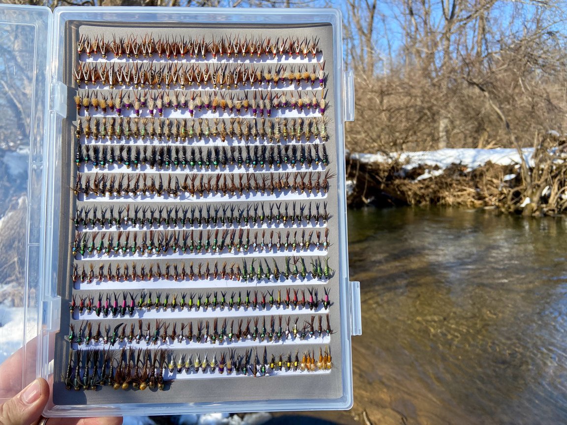 A collection of small flies held up in front of a wintery trout stream.