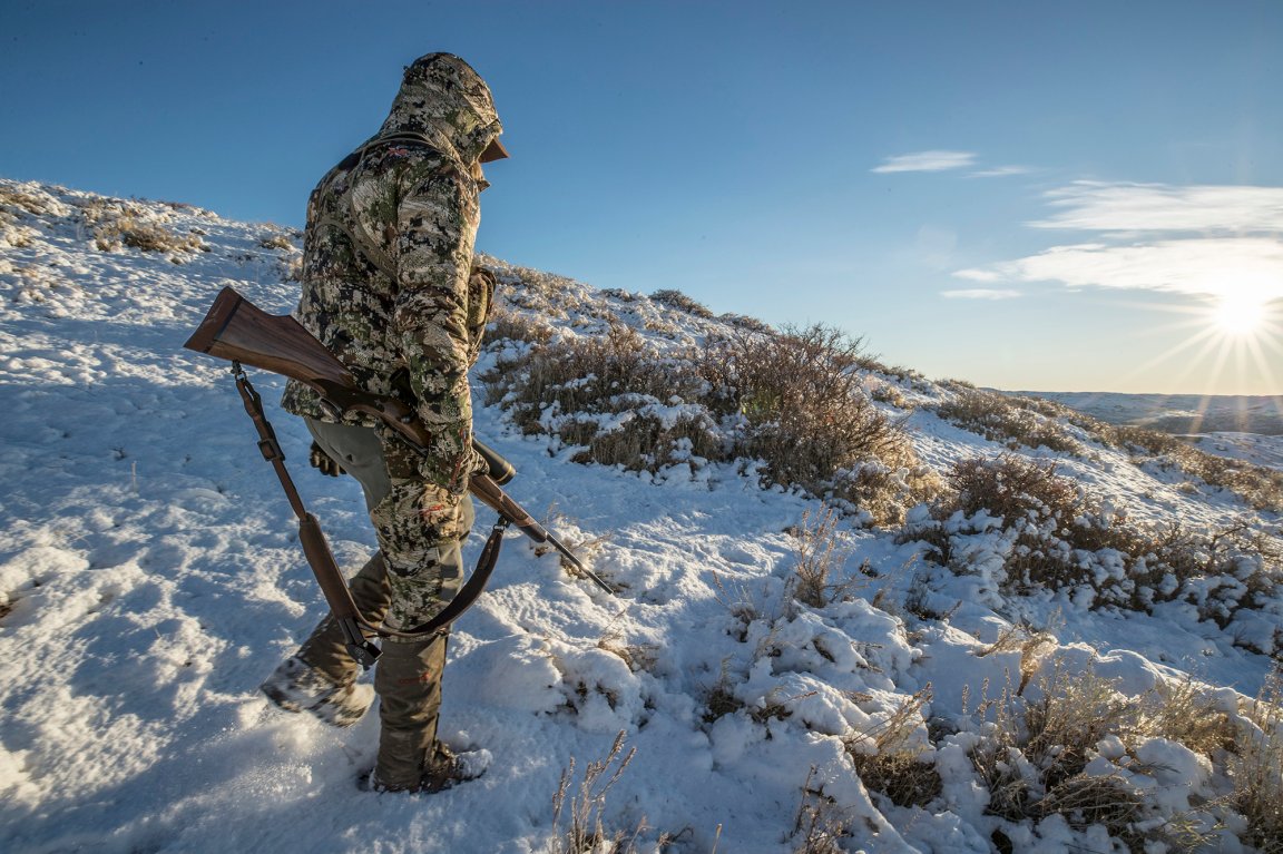 A hunter in head-to-toe sitka camo carries a wooden rifle.