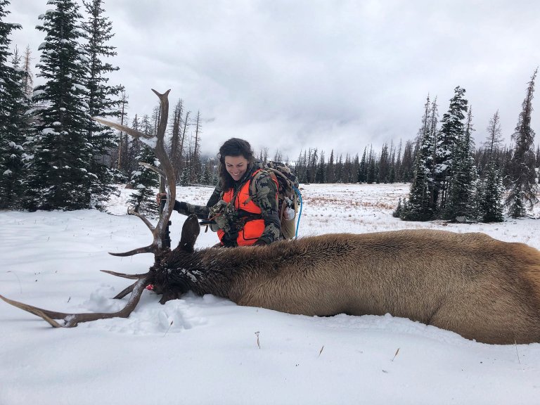 A hunter in kuiu and an orange vest sits behind a bull elk in the snow.