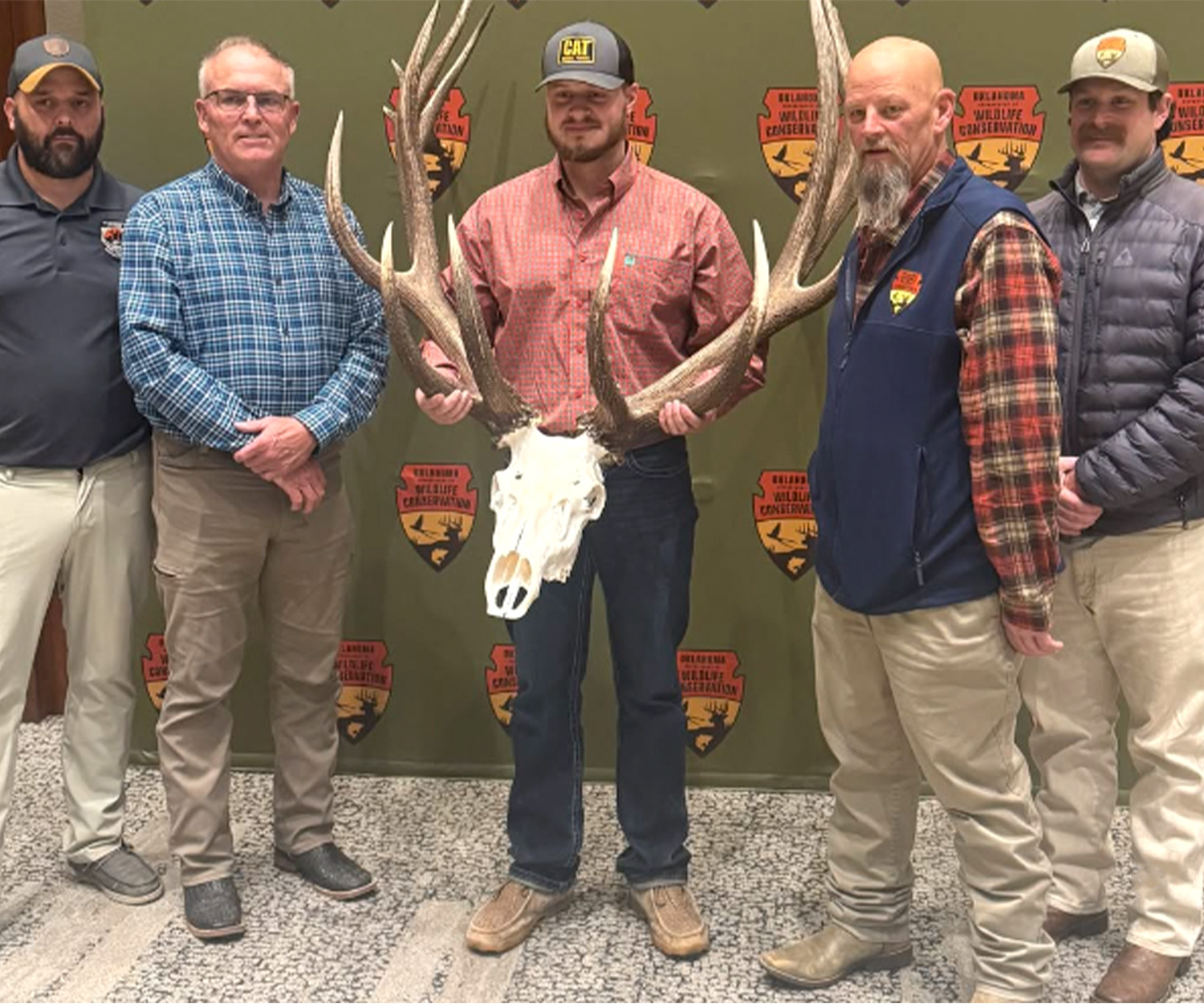 Five men stand in front of a step and repeat with the Oklahoma wildlife conservation logo and the giant nontypical bull.