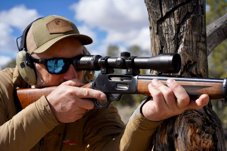 A photo of a John Snow shooting a wooden-stocked lever action off a tree.