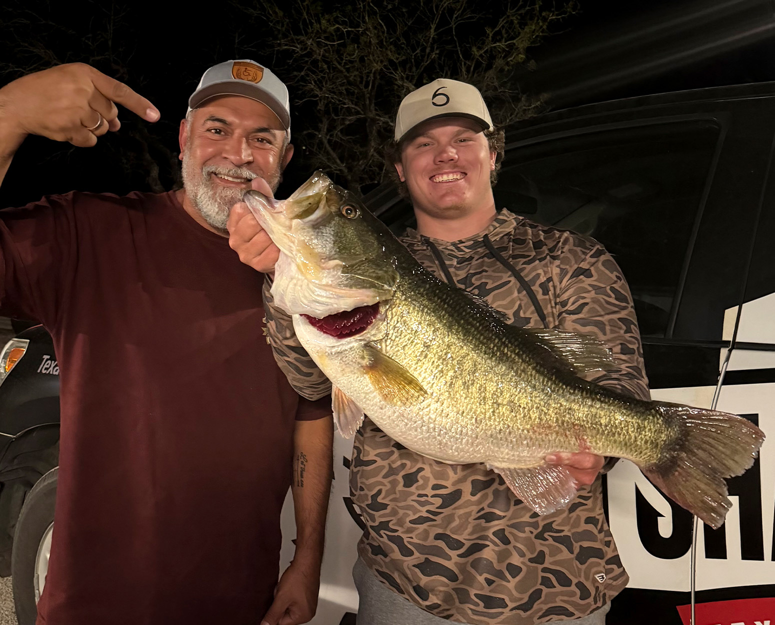 A fisherman holds up a big bass while the guy next to him points to it.