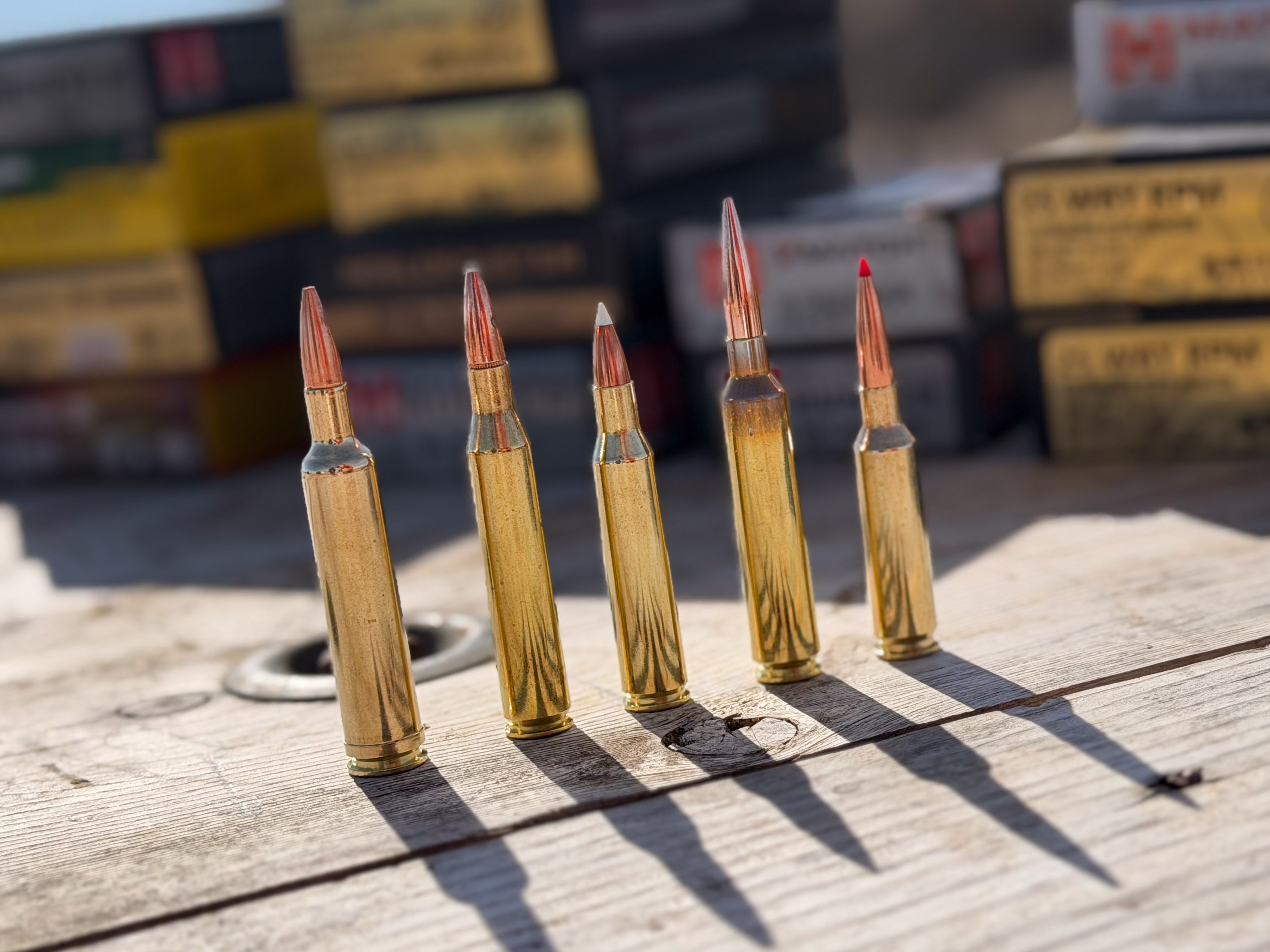 Five rifle cartridges in a row on a table of ammo. From .257 Weatherby Magnum, .25/06 Remington, .257 Roberts, 25 Weatherby RPM, and the 25 Creedmoor.
