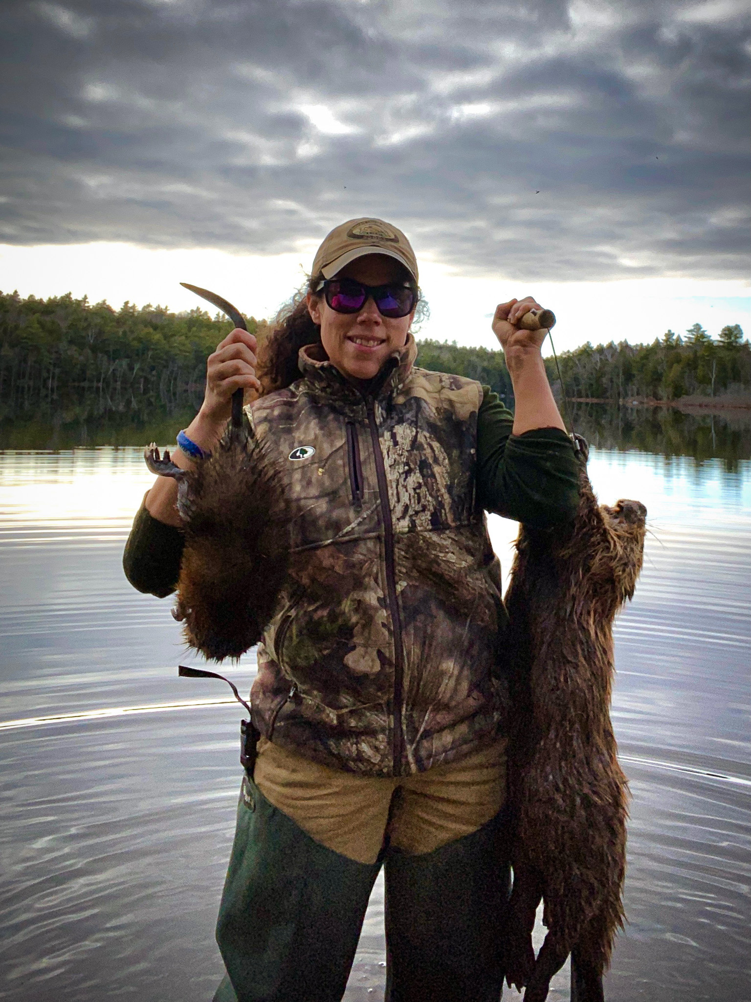 A woman in camo holds up a wild-trapped muskrat and a beaver.