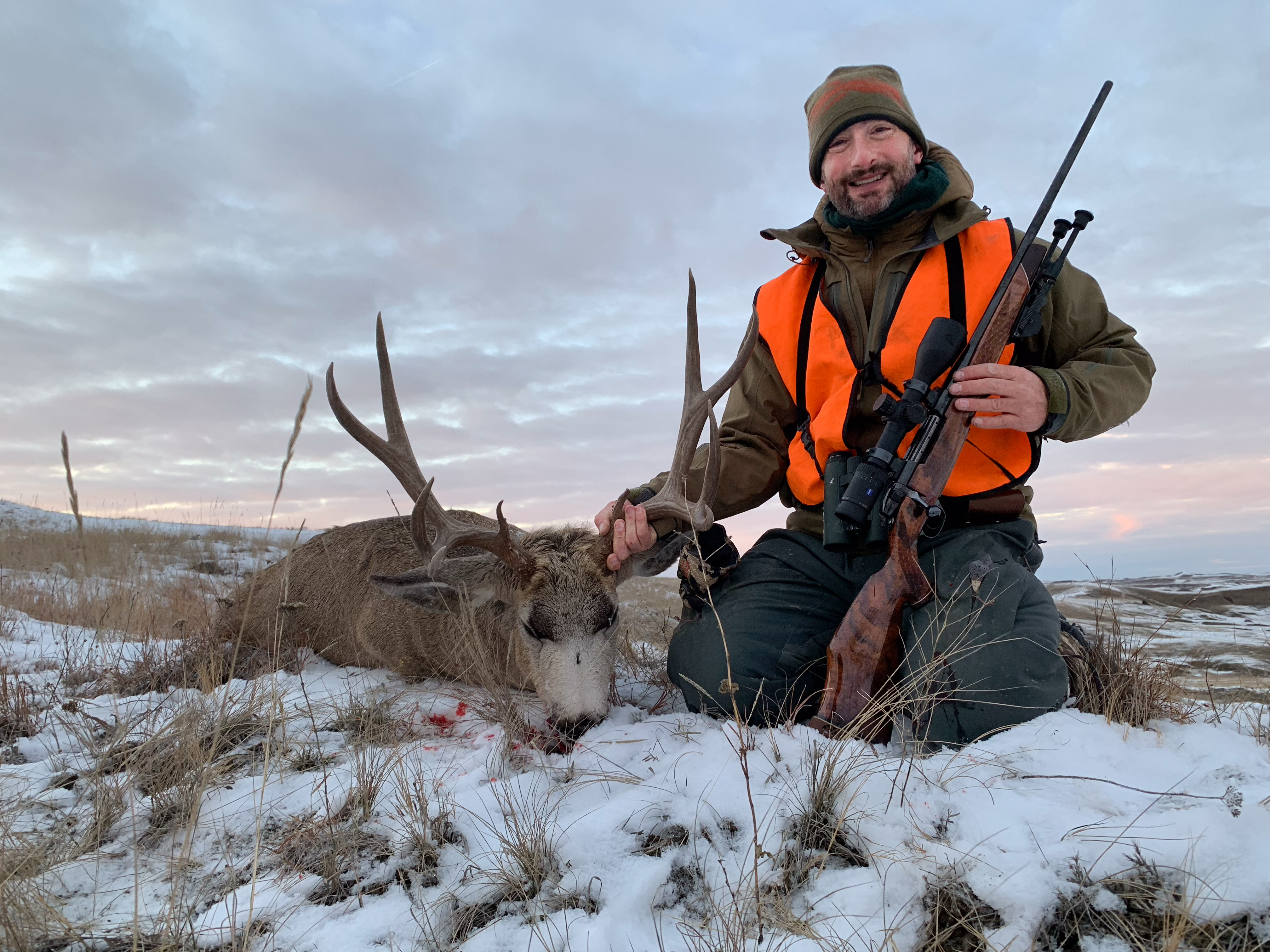 A hunter in blaze orange kneeling next to a mule deer buck in the snow while holding a wooden .257 weatherby magnum rifle