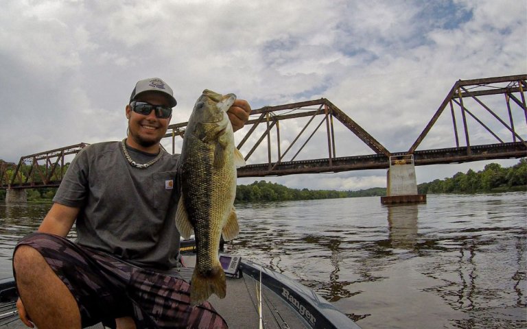 An angler holds up a large spotted bass in front of a bridge.